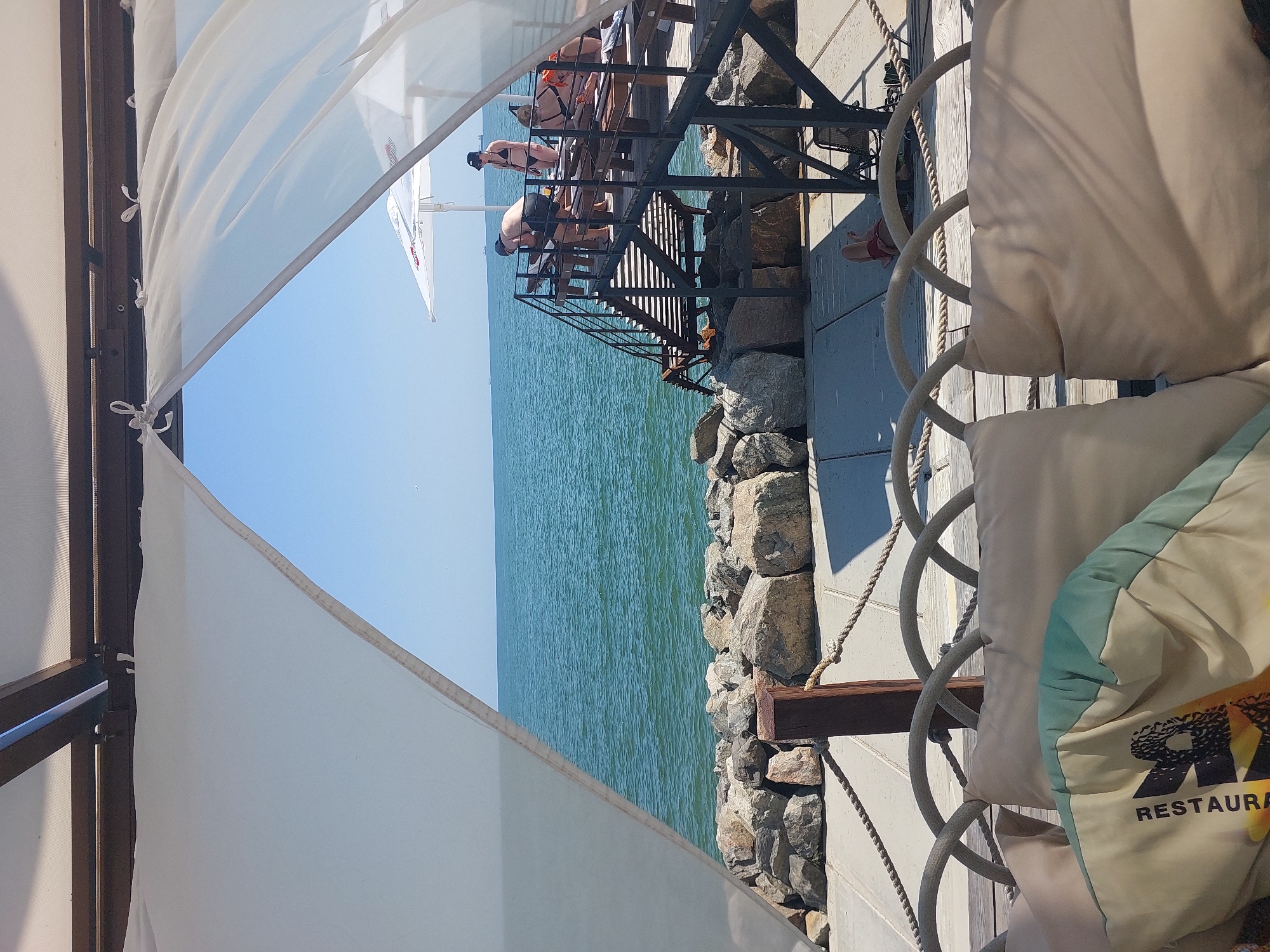 The image shows a scene with people near a body of water. The perspective is from underneath a white canopy, looking out towards the water. In the mid-ground, there are several people on a metal staircase that leads down towards the water. Some of the people appear to be in swimsuits, suggesting a recreational setting. The water is a vibrant blue-green and appears calm, with small ripples on the surface. The staircase is next to a rocky embankment. In the foreground, to the right, there are large, light-colored cushions, possibly part of outdoor seating. A rope railing is visible, intertwined with a metal structure.
The time of day appears to be daytime, with bright sunlight casting shadows. The sky is a clear blue, indicating fair weather. The overall impression is one of a leisurely day at a waterfront location, possibly a restaurant or beach resort.
On one of the cushions in the foreground, there is visible text that reads "RESTAURA". The word is part of a larger logo or design that includes the letters "RA" in a bold font and an image that appears to incorporate sunflowers. The setting seems to be outdoors, with the natural elements of water and rocks juxtaposed with man-made structures like the canopy and staircase.