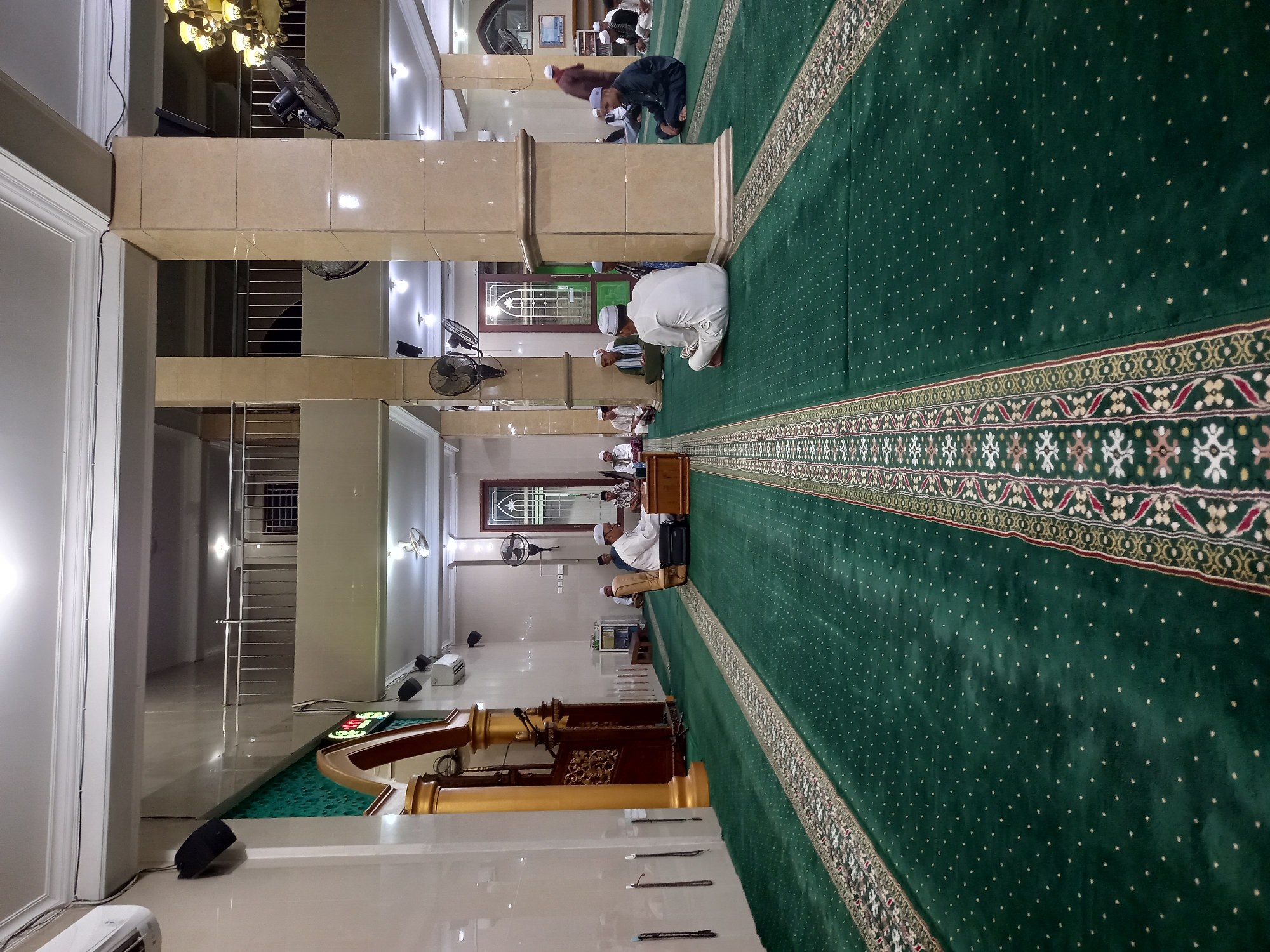 The image depicts the interior of a mosque, likely in Samarinda, Indonesia, judging by the presence of a digital clock displaying the time in the local format and the architectural style. The floor is covered with a plush, dark green carpet featuring intricate patterns along the aisles, indicative of a prayer rug. Several men, dressed in traditional Islamic attire such as sarongs and peci (caps), are seated on the carpet, facing various directions. Some appear to be engaged in prayer or religious discussion, while others are observing.
In the foreground, a man in a white outfit with a white peci is seated cross-legged, facing away from the camera. To his left, another man in a dark outfit and white peci is also seated cross-legged. Towards the center of the image, a group of men are gathered around a wooden podium. One man, in a white robe and peci, is seated at the podium, possibly leading a prayer or delivering a sermon. Another man, in a dark robe, is standing and gesturing.
The mosque's interior is characterized by large, square pillars with marble-like cladding. The walls are painted in light, neutral tones, and there are numerous wall-mounted fans, indicating a warm climate. Decorative wooden elements, including what appears to be a minbar (pulpit), are visible on the left side of the frame. The lighting is a mix of recessed ceiling lights and chandeliers, creating a well-lit environment. The presence of air conditioning units mounted on the walls suggests a modern facility. The overall atmosphere is one of solemnity and communal gathering.