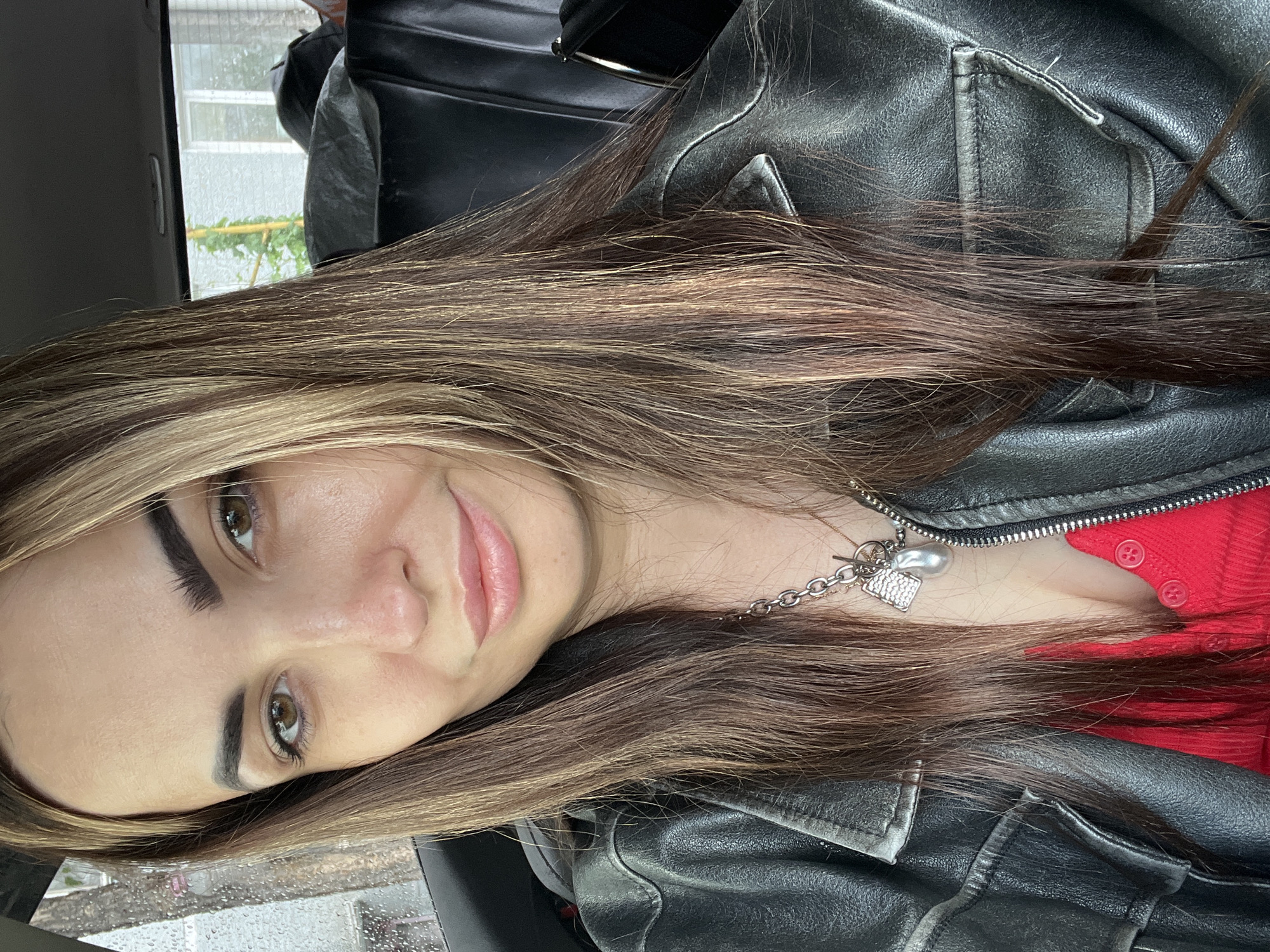The image displays a portrait of a young woman, originally oriented in a landscape format and rotated 90 degrees clockwise. When viewed upright, she is looking directly at the camera with a soft, subtle smile. She has long, straight brown hair with noticeable blonde highlights, framing her face and resting over her shoulders. She is dressed in a black leather jacket, partially unzipped, revealing a bright red top underneath with small buttons visible down the front. Around her neck, she wears a silver-colored chain necklace adorned with multiple charms, including a distinctive teardrop-shaped pearl and a smaller, possibly rectangular, shiny pendant. Her facial features include well-defined dark eyebrows, brown eyes, and light pink lips.
The setting appears to be inside a vehicle, likely a car. Through a side window in the background, numerous water droplets are clearly visible on the glass, indicating that it is raining or has recently rained. Beyond the wet window, a blurred view of a building and some green foliage suggests an urban or suburban outdoor environment. Parts of the vehicle's dark interior, possibly the back of a seat, are also visible. The lighting in the image suggests it is daytime, although the rainy weather might imply an overcast sky. The woman's expression conveys a calm and pleasant mood. There is no visible text in the image.