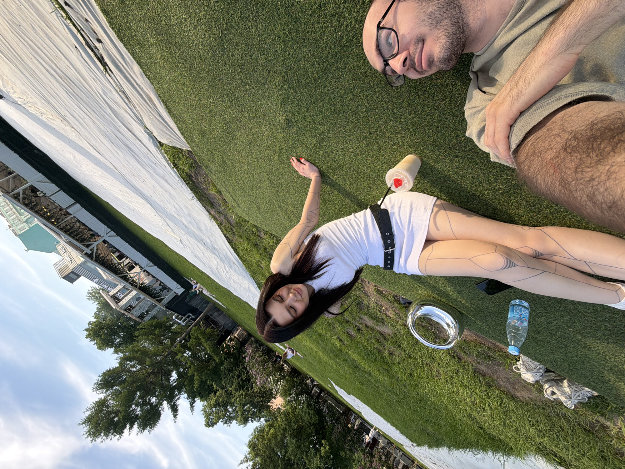 The image shows a grassy area outdoors. A woman lies on the grass wearing a white dress and black belt, with black ink tattoo-like designs on her legs and arm. A man lies next to her, also on the grass. Next to the woman are a metal bowl, a water bottle, and a pair of sneakers. A drink is next to the man. A greenhouse-like structure is visible in the background. Trees and a blue sky with some clouds are also visible in the distance. Based on the greenery and building structure, this could be in Moscow, Russia.