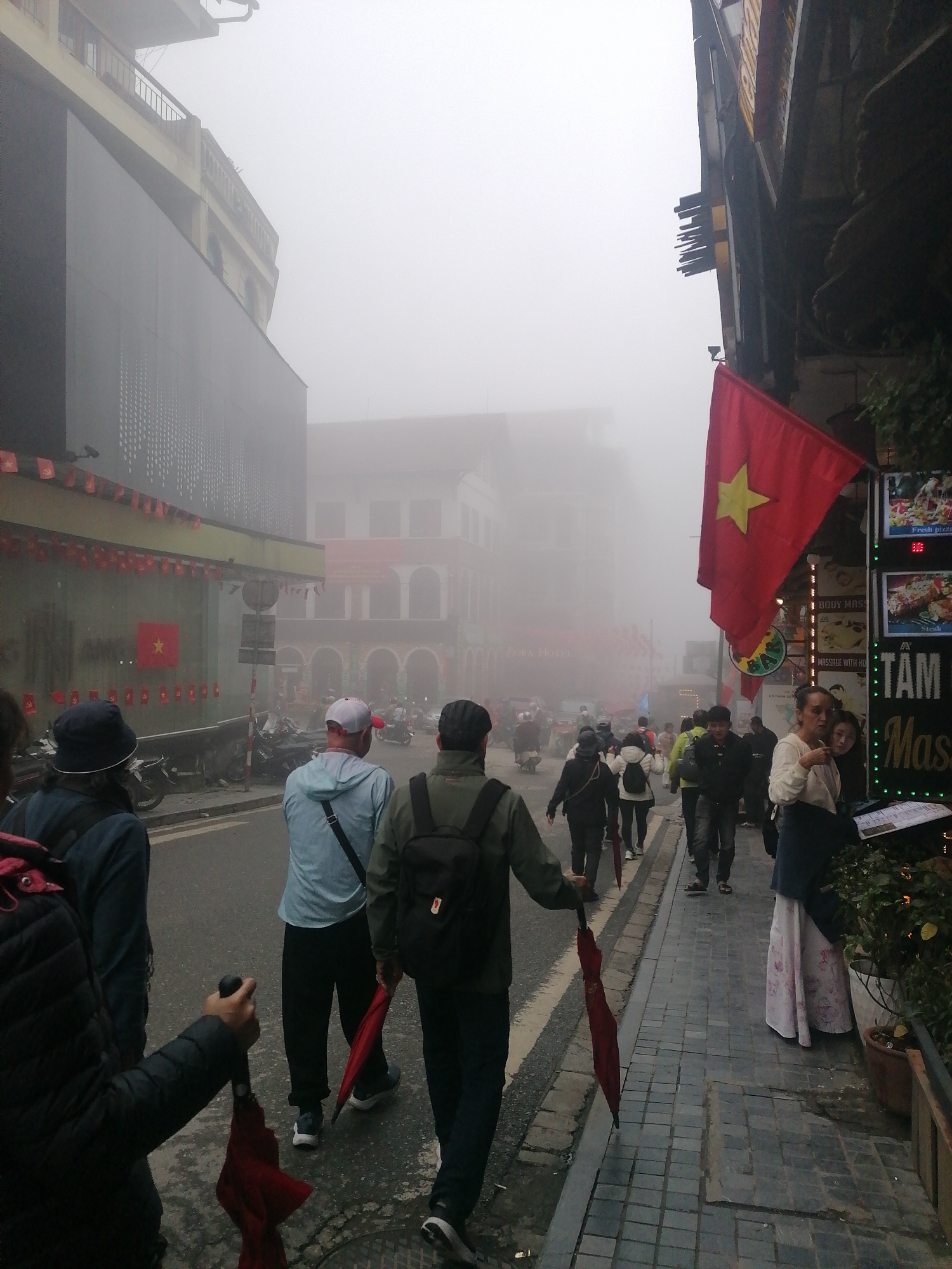 The image depicts a busy street scene in Sa Pa Ward, Vietnam, enveloped in thick fog, creating a soft and somewhat obscured atmosphere. The dense mist limits visibility of background buildings, giving them a blurred, ethereal quality.
Numerous pedestrians are walking along the street and sidewalks. In the foreground, several individuals are walking away from the camera; one person on the far left holds a closed red umbrella, and another in the middle also carries a red umbrella. People are generally dressed in jackets, suggesting cool weather. Motorbikes are parked along the left side of the street, indicating common urban transportation.
On the left, a large building with a dark, textured facade is decorated with several small red banners bearing yellow stars, consistent with Vietnamese flags. Further down the street, another larger red flag with a prominent yellow star hangs vertically from a building on the right side. Below this flag, a woman in a long, patterned skirt stands near an illuminated menu display or sign. The signs visible on the right advertise services such as "Fresh pizza," "Body Massage," "Steak," and "TÀM Massage," with partially visible text.
The scene portrays daily life in Sa Pa under foggy conditions, with people moving purposefully through the town. The overall impression is one of a bustling local area, possibly catering to tourists, given the nature of the advertisements. The fog is the most striking weather detail, transforming the visual clarity of the environment.