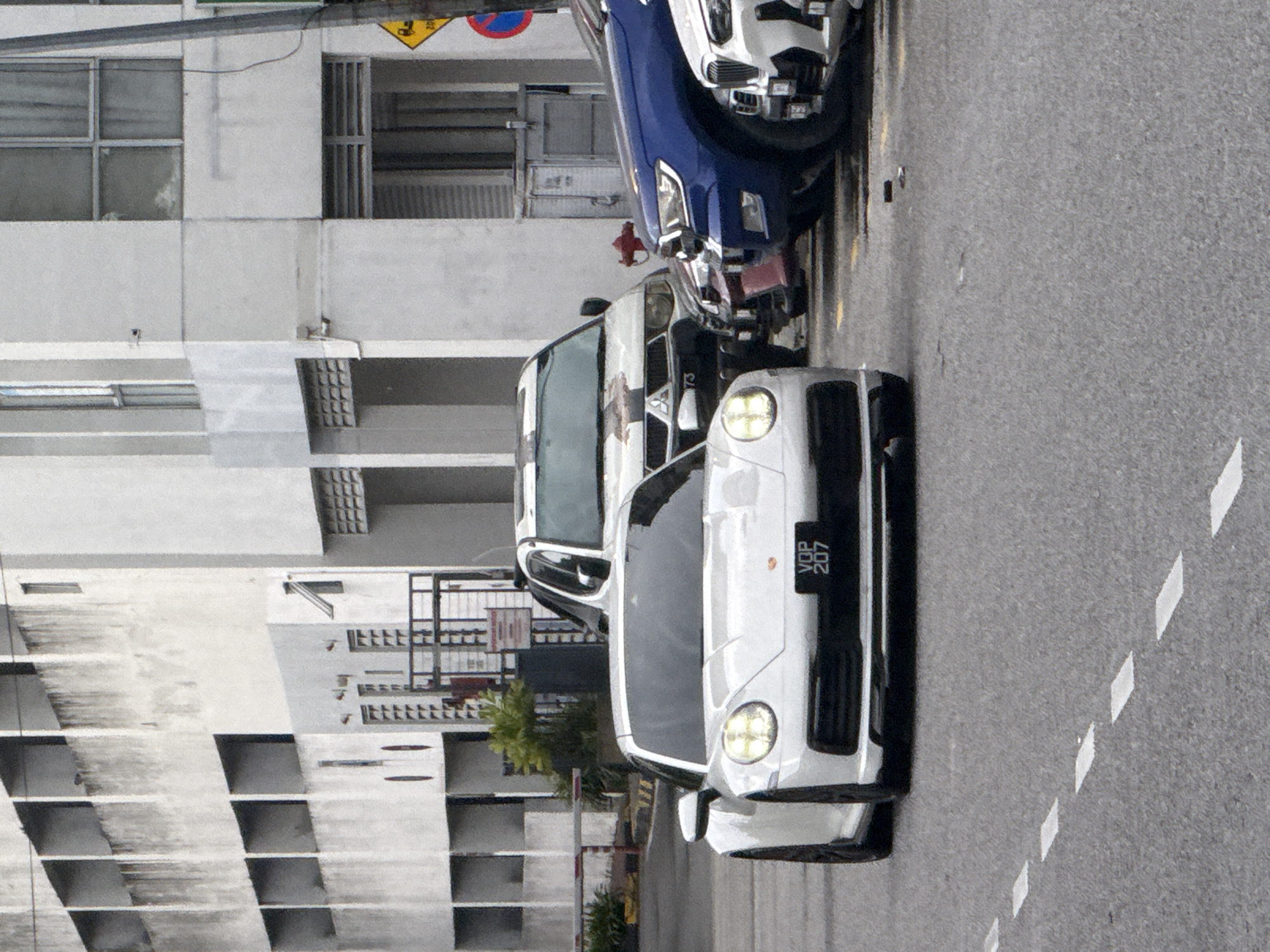 The image depicts a street scene in Ampang Jaya Municipal Council, Malaysia. Three vehicles are visible: a white Porsche with license plate VDP 207 in the foreground, followed by a white van, and a dark blue SUV. All cars appear to be parked on the side of the road. A multi-story building with a repeating pattern of windows and cubicles dominates the left side of the frame. Street signs are partially visible above the vehicles. The road has white dashed lines.