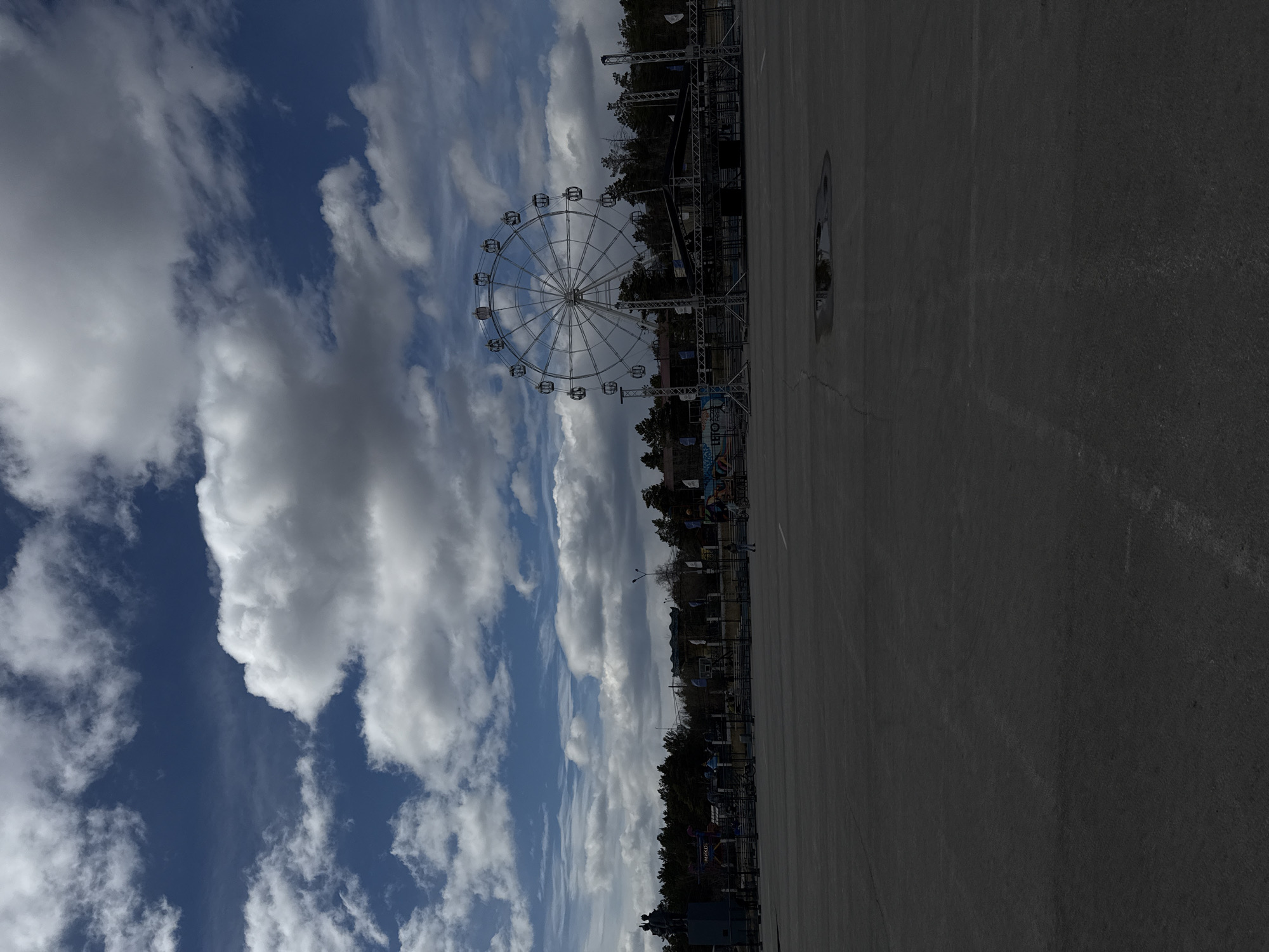 The image depicts a Ferris wheel in an outdoor area of Yakutsk, Russia. The Ferris wheel is white and features enclosed cabins. The sky is blue with numerous white, puffy clouds. Adjacent to the Ferris wheel is a paved area, and structures resembling scaffolding or event staging are visible. A line of trees borders the back of the area.