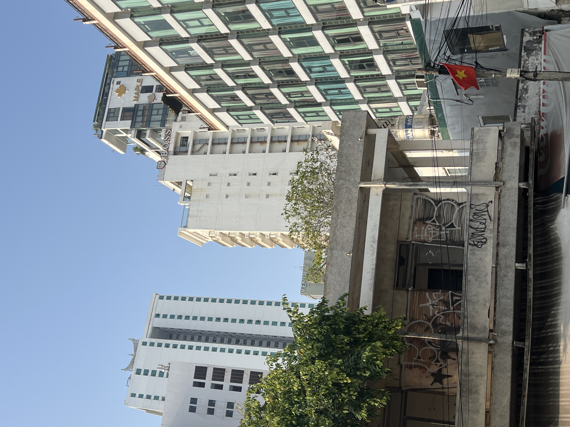 The image shows a street scene in Nha Trang, Vietnam, looking up at buildings. There are several tall buildings in the background, mostly white, with rows of windows. One building is a taller, modern glass-windowed structure. An older, deteriorating building is in the foreground, with graffiti on its exterior walls. A Vietnamese flag is visible, partially obscured by electrical wires. Green foliage from trees or bushes partially obscures the lower portion of the building. The sky is blue.