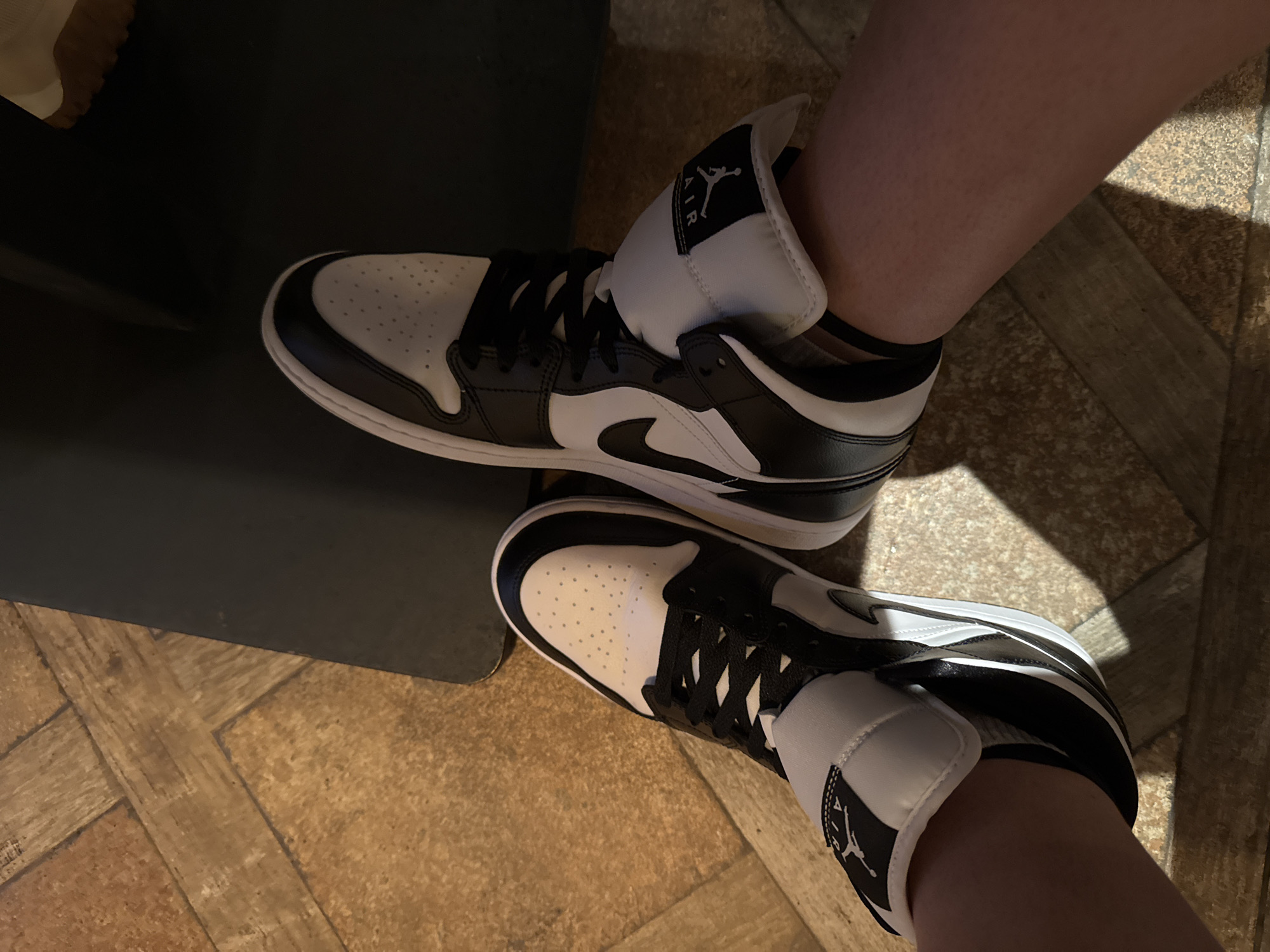 A pair of black and white high-top sneakers, appearing to be Air Jordan 1s, are worn on a person's feet. The sneakers have black laces, a black swoosh logo, and white toe boxes with perforations. The person's legs and feet are visible, resting on a floor made of wooden planks. The floor has a parquet pattern and a warm, brown tone. A dark, solid object, possibly a piece of furniture or a wall, is partially visible in the background. The lighting suggests an indoor environment, with shadows cast on the floor by the sneakers. The scene does not contain any identifiable landmarks or context to suggest a specific location in Varna, Bulgaria.