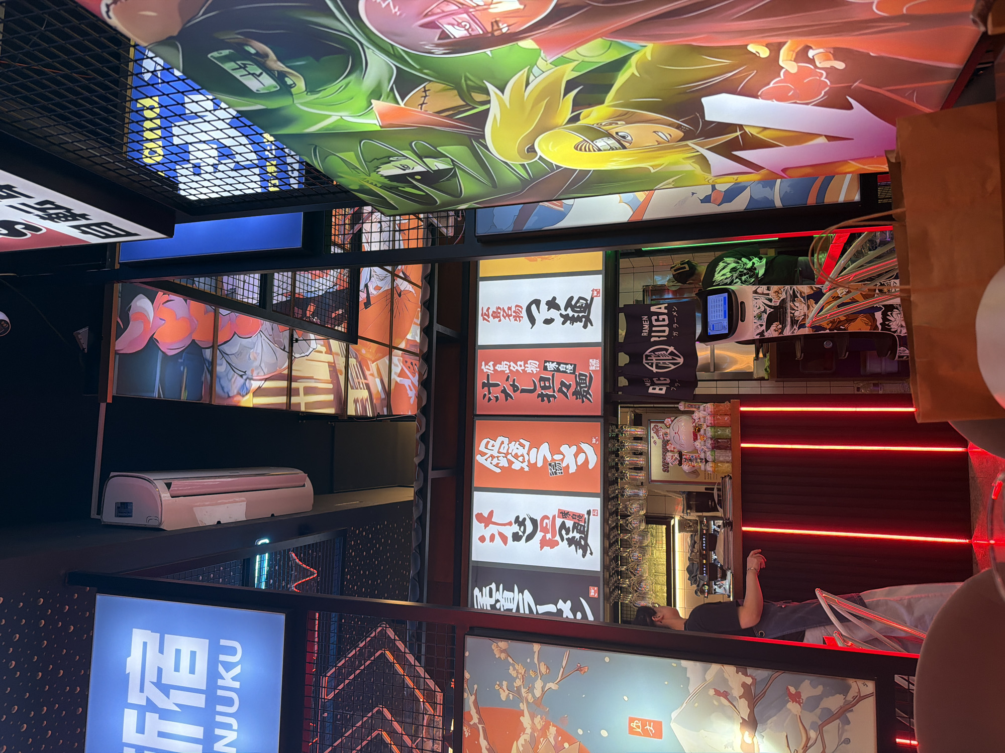 The image depicts the interior of a ramen restaurant, likely in Lisbon, Portugal, based on the signage and style of the establishment. Dominating the right side of the frame is a large, vibrant mural featuring characters from the anime series "Naruto." To the left, illuminated signs with Japanese text advertise different types of ramen. In the center, a bar area is visible with a person behind it, seemingly preparing drinks or taking orders. The bar is decorated with red neon lights. Various bottles of liquor and glasses are stocked on shelves above the bar. An air conditioning unit is mounted on the wall near the top left. The overall atmosphere is modern and visually engaging, with a strong theme of Japanese culture and anime.