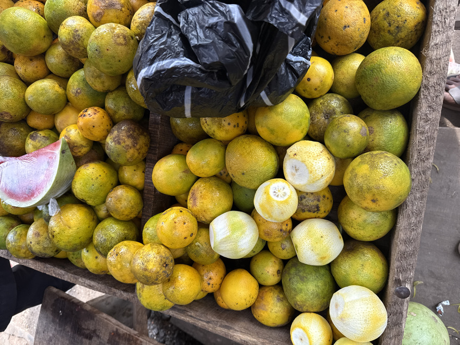 This image depicts a market stall in Ogere, Nigeria, overflowing with a large quantity of citrus fruits, primarily oranges. The oranges display various shades of yellow, green-yellow, and orange, many exhibiting natural blemishes and dark speckles. Several oranges appear partially peeled or cut, exposing white pith. A slice of watermelon, featuring pink flesh and dark seeds, wrapped in clear plastic, rests atop a pile of oranges near the upper center. A crumpled black plastic bag with white horizontal stripes is situated on the right side, partially covering some fruits. The fruits are contained within a rustic, weathered wooden structure, likely a crate or display stand. In the upper left corner, a dark-clothed individual is partially visible, while at the bottom, light-colored footwear suggests the presence of another person near the stall. A large, green-skinned fruit, possibly a watermelon, is partially visible at the bottom left.