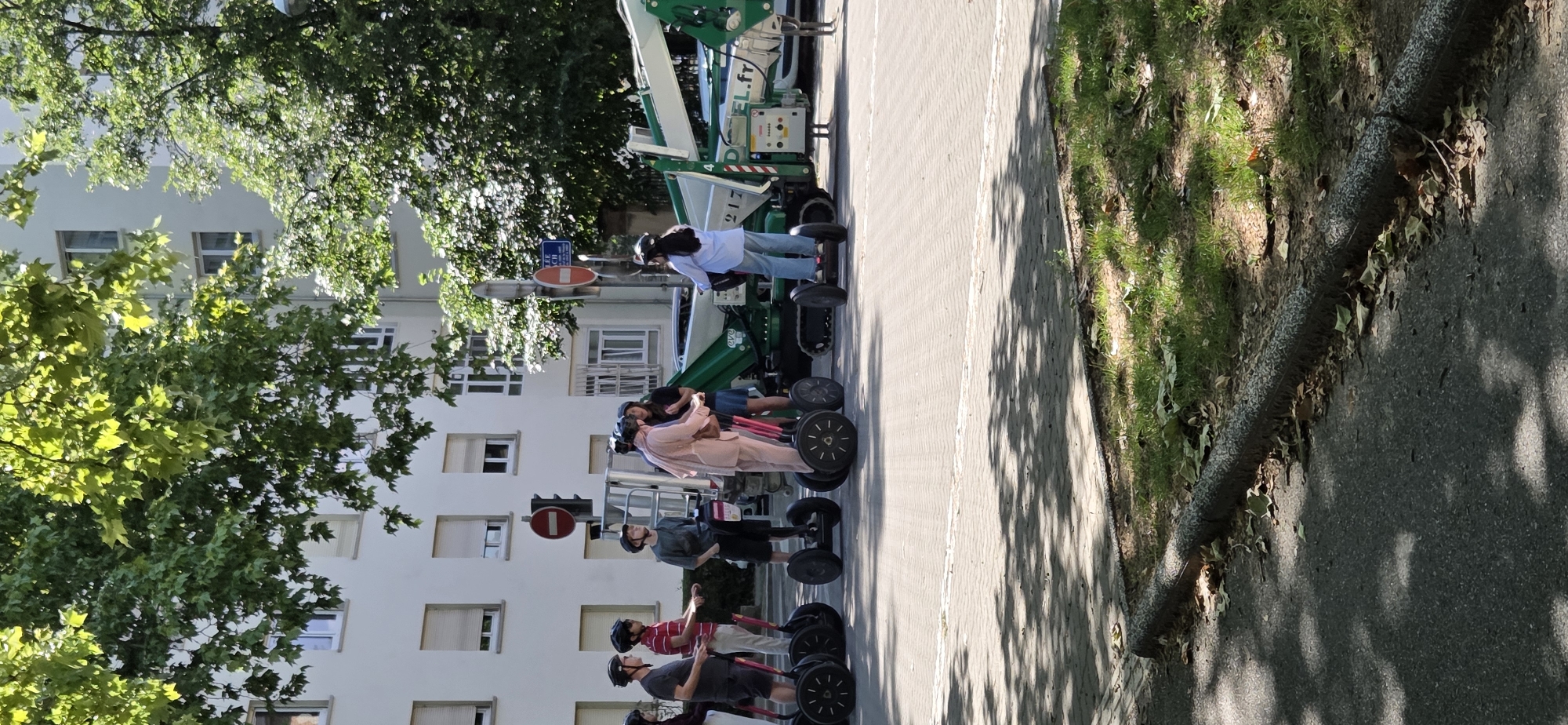 A group of people on segways are gathered near a building and a crane in what appears to be an urban environment. The people are dressed casually, and some are wearing helmets, suggesting a recreational activity or tour. One person in a light blue shirt is standing next to a segway, while others are on their segways, some interacting with each other. The building in the background is multi-story with numerous windows. A green crane is visible in the background, possibly involved in construction or maintenance work. The scene is illuminated by daylight, and the presence of shadows suggests it is likely daytime. The overall atmosphere seems relaxed and social. There is some visible text on the crane, including "217" and "EL.fr". There are also traffic signs, one red and white circular sign with a horizontal bar, and another smaller sign above it. The ground is paved with cobblestones in some areas and asphalt in others, with a grassy area bordering the paved path. The image appears to be taken outdoors on a sunny day.