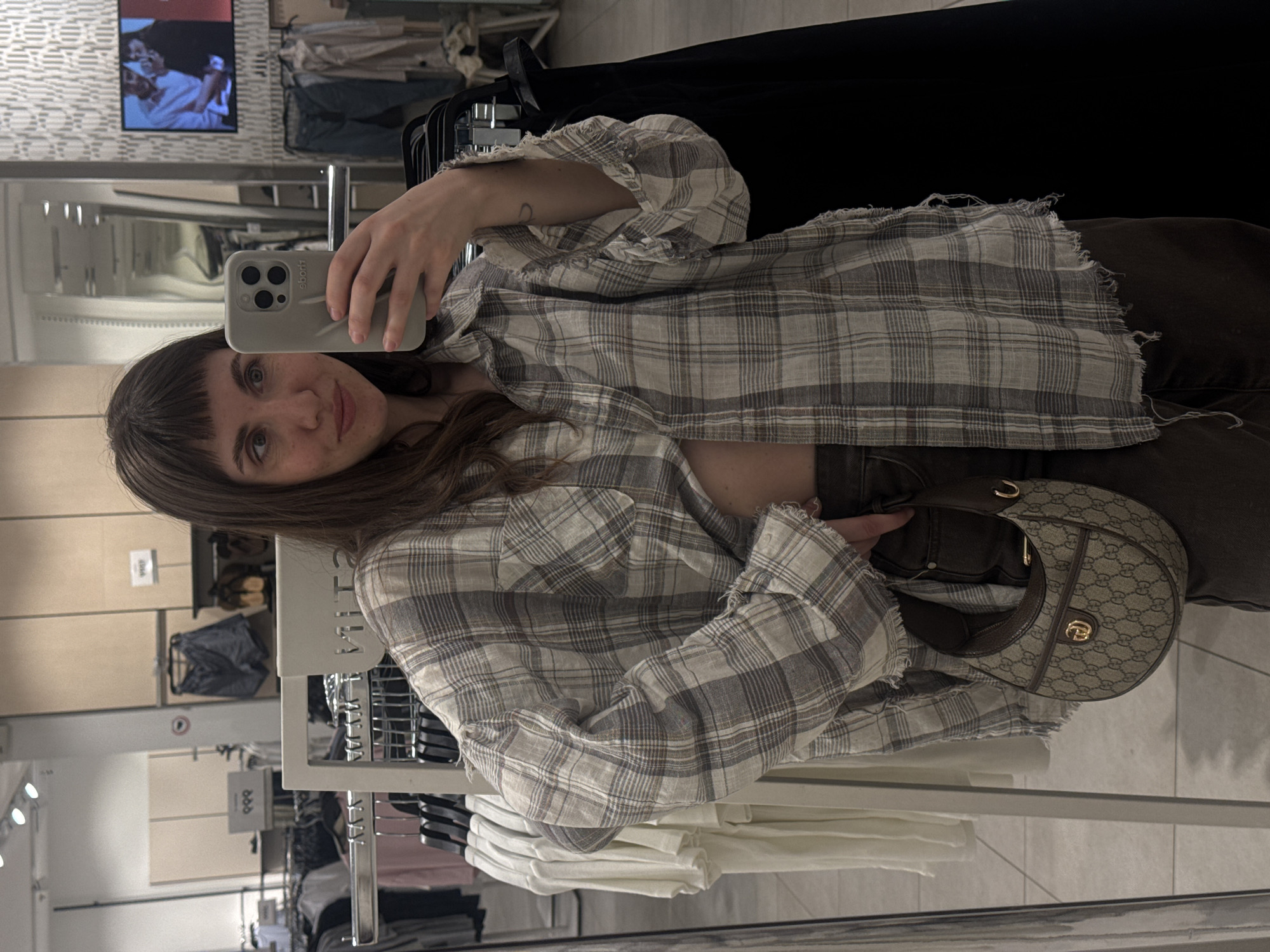 A woman is taking a selfie in what appears to be a clothing store dressing room. She is holding her phone in her left hand, with a gray phone case visible. The woman is wearing a gray and white plaid shirt and dark-colored pants or jeans. She also has a patterned handbag with a gold emblem.
In the background, there are clothing racks filled with various items of clothing. There's also a television screen showing a picture, and the reflection of the clothing store. It is not possible to definitively identify if this is Moscow, Russia.
