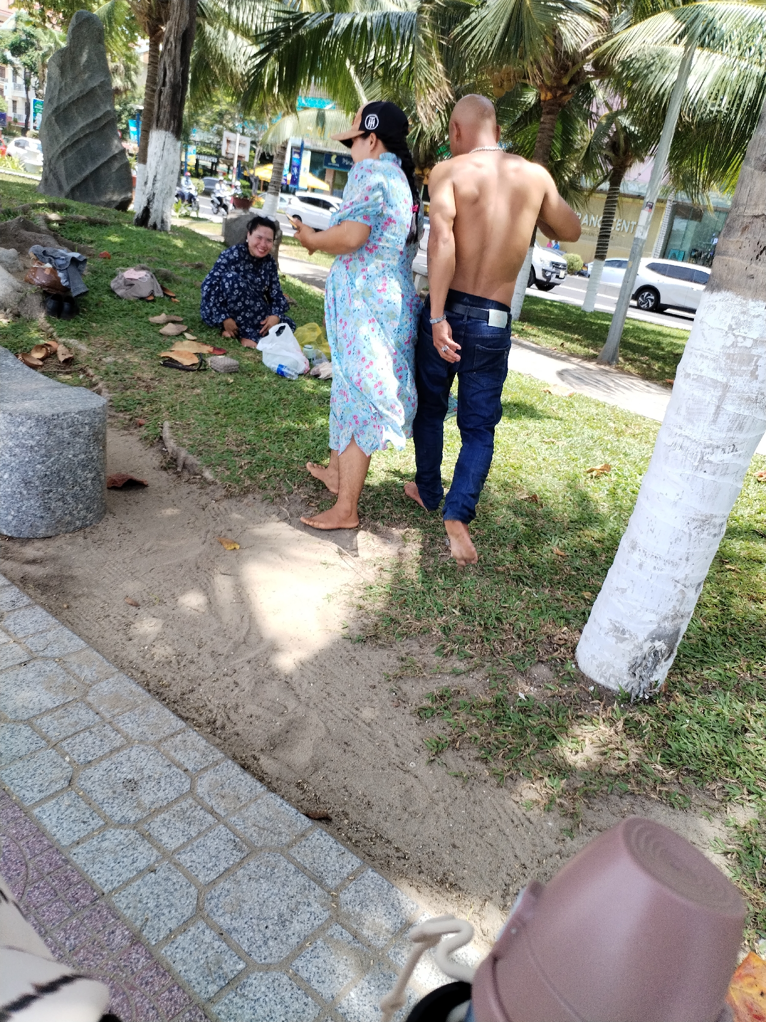 The image depicts an outdoor scene on a sunny day in Nha Trang Ward, Vietnam. In the foreground, a person wearing a floral-patterned dress and a black cap is interacting with another person, a bare-chested man in blue jeans. The woman is holding up a smartphone, seemingly taking a photo or video of the man, who is facing away from the camera with his back to the viewer. The man has a shaved head and is wearing a necklace.
The two individuals are standing on a grassy area next to a sandy path. Behind them, there are palm trees and some buildings visible in the background, suggesting an urban or resort setting. On the grass to the left, a person in a dark blue patterned garment is seated, looking towards the other two individuals. Scattered around the seated person are various items, including a white plastic bag, some footwear, and what appear to be pieces of food or wrappers.
To the left of the frame, a large, grey, textured concrete structure is visible, possibly a bench or a decorative element. The ground transitions from a paved area with a patterned design to sand and then grass. The overall atmosphere suggests a casual, public gathering or leisure activity.