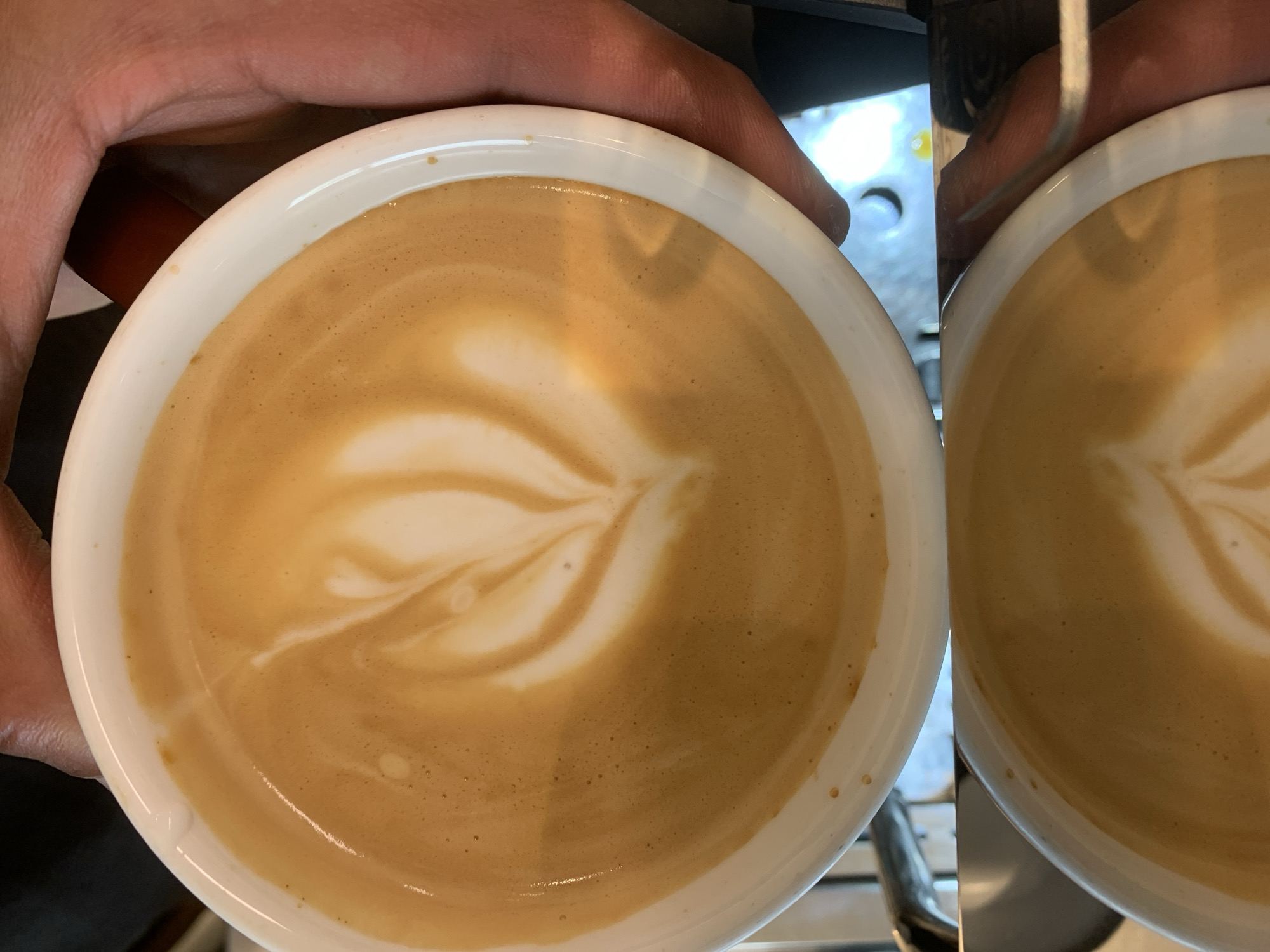 A high-angle, close-up shot shows a hand holding a white, circular cup filled with a tan-colored beverage, likely coffee with milk, exhibiting latte art in the form of a leaf-like pattern. A second similar cup is partially visible to the right of the main cup. The cups are sitting on a metal surface, possibly part of a coffee machine. The lighting suggests an indoor environment. No other details suggest Barcelona, Spain.