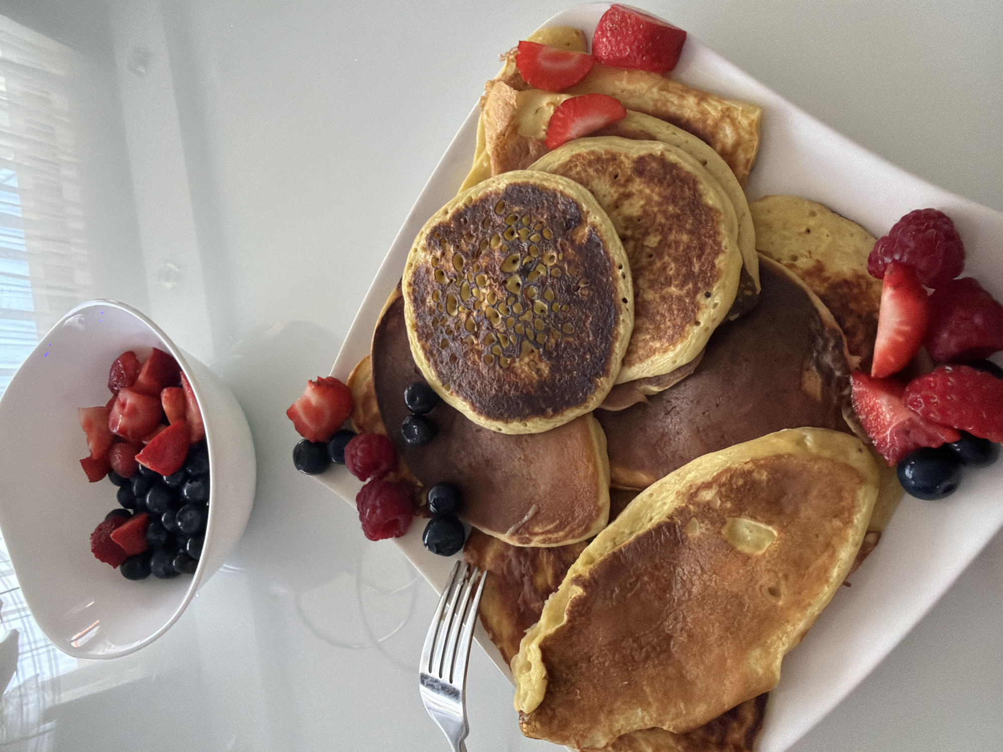 The image shows a stack of pancakes and some crepes on a white plate, accompanied by a white bowl of mixed berries (strawberries and blueberries). Some additional strawberries, blueberries, and raspberries are arranged around the edges of the pancakes. A silver fork is also visible on the plate. The pancakes vary in color and texture, with some appearing darker and others more golden.
There's no information to suggest this image was taken in Gabrovo, Bulgaria.