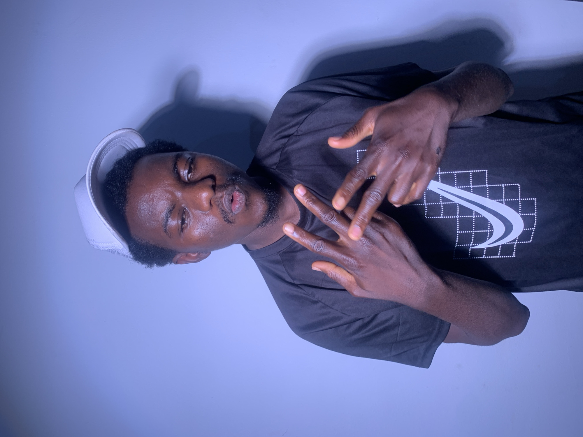 A young Black man, wearing a white baseball cap and a black t-shirt with a white Nike logo, is photographed from the waist up. He has dark skin, short black hair, and a goatee. His head is tilted to the right, and his eyes are looking toward the camera. He is making a "peace sign" gesture with his right hand, with his index and middle fingers extended, and his thumb, ring finger, and pinky finger curled. His left hand is partially visible, also held up in a gesture. The background is a solid, pale blue, with a shadow of the man cast on the wall behind him, suggesting a light source from the left. The lighting appears to be artificial, possibly from a studio setting. The image is vertically oriented and appears to be taken indoors. The overall mood is casual and somewhat playful. There is no visible text other than the Nike logo on his shirt.