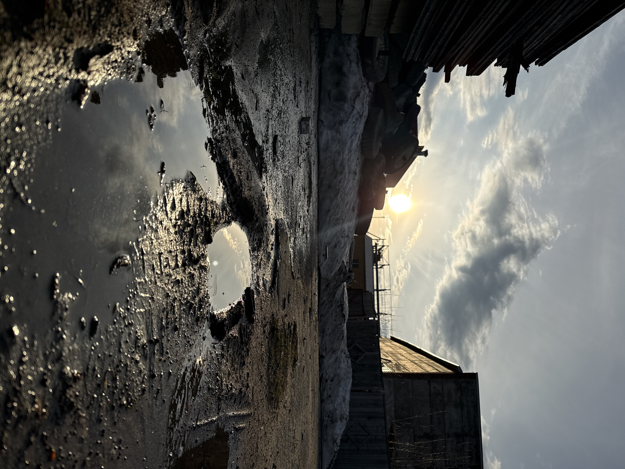 The image depicts an urban scene, likely in Chaikovsky, Russia. A large puddle occupies the foreground, reflecting the sky and clouds. The ground around the puddle is muddy and uneven. To the right of the puddle is a rough, possibly unpaved area adjacent to a snow drift. Beyond the snow drift are various buildings or structures, including what appear to be a wooden shed or warehouse and a taller building with antenna. The sun is positioned in the upper center-right, creating a bright glare and silhouetting objects. Debris or construction materials, possibly wooden planks, are piled along the top edge of the image. The sky is partially cloudy. There are no visible people or explicit interactions taking place.