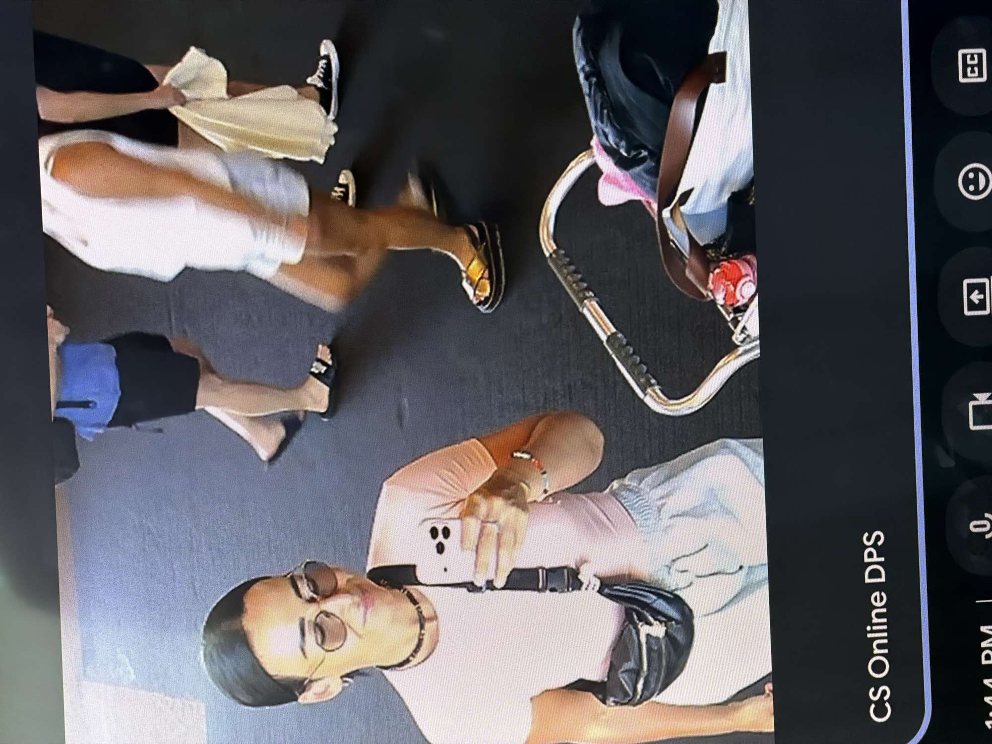 The image shows an overhead view of a group of people and objects on what appears to be a flat, gray surface, possibly a street or sidewalk. One individual, wearing sunglasses, a light pink shirt, and light blue shorts, is taking a photograph with a white smartphone. They are also wearing a black fanny pack across their chest. Other individuals are visible walking in different directions. One person wears black shorts with a blue accent, another wears a white outfit with gold sandals, and a third wears black shoes. A baby stroller is also present. Based on the prompt, this location is assumed to be in Kuta, Indonesia.