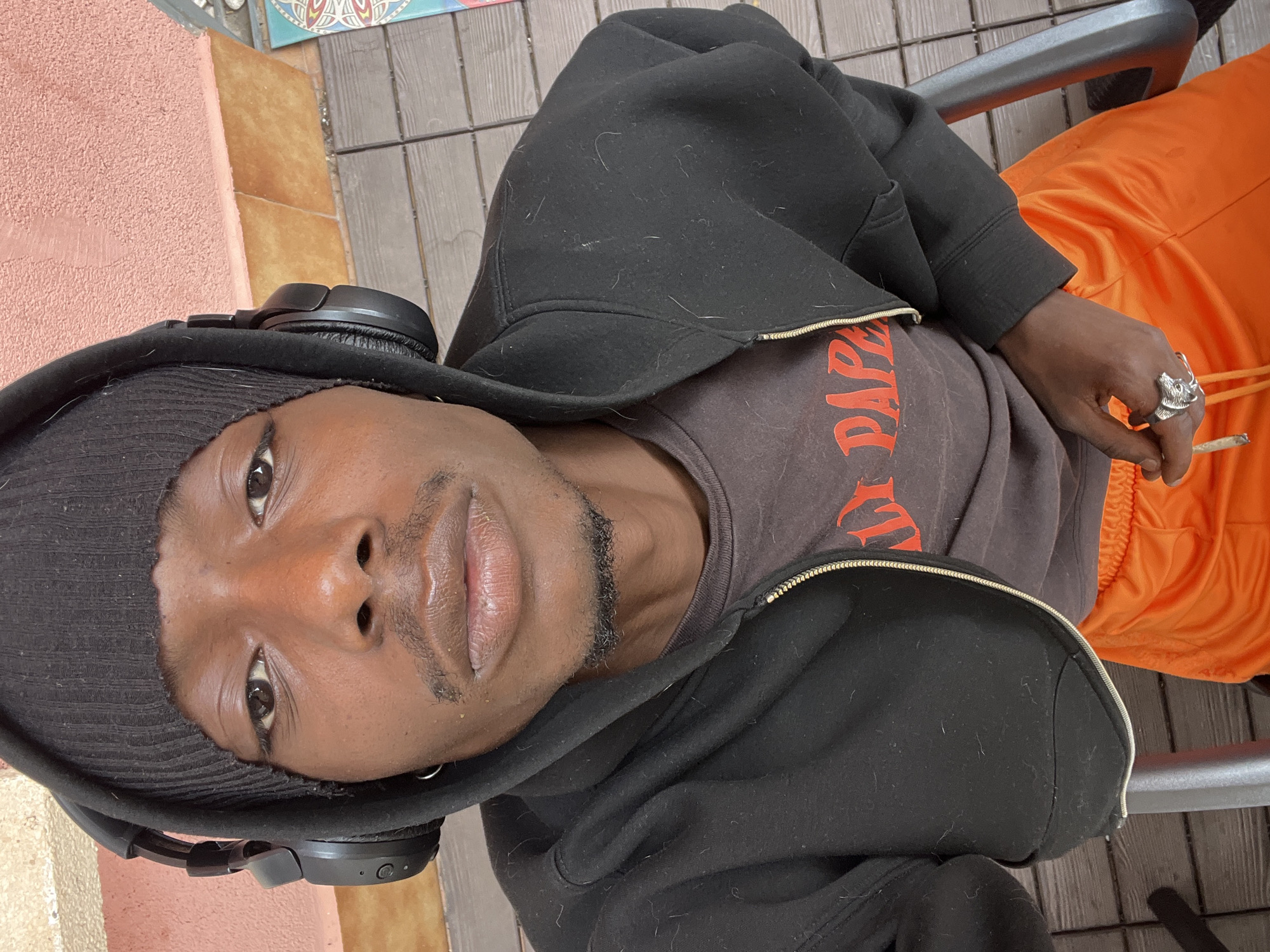 A young man with dark skin and a short stubble beard and mustache is captured in a medium close-up, looking directly at the camera. He wears a black ribbed beanie, black over-ear headphones, a black zippered hoodie (which appears to have lint or fibers), and a dark gray t-shirt with orange text. The visible text on his shirt appears to read "PAPERS". He is also wearing bright orange track pants. On the ring finger of his right hand, he sports a prominent silver-toned ring shaped like an animal's head. He holds a hand-rolled cigarette or joint between the thumb and forefinger of his right hand.
The setting appears to be an outdoor patio or balcony in Barcelona, Spain, with a light pink textured wall above terracotta-colored tiles, and dark wooden plank flooring. A colorful decorative tile or artwork is partially visible in the upper background. The bright, even lighting suggests it is daytime. The man has a neutral, direct gaze. He is seated in a chair, with a gray metal chair arm visible to his right.