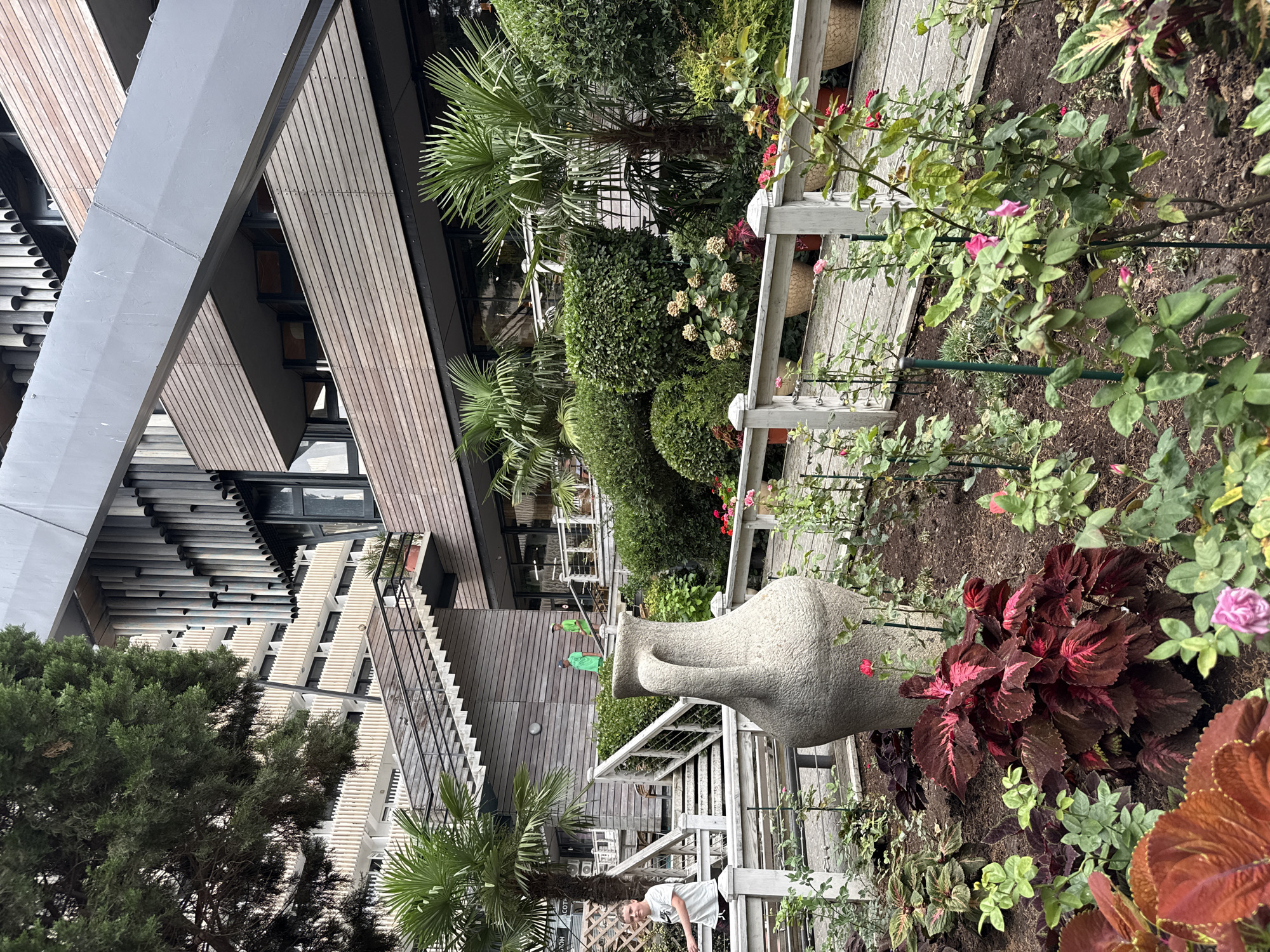 An exterior shot captures a garden area adjacent to a multi-story building with a modern architectural design. The building features horizontal wooden cladding and angled metallic structures. In the foreground, a large, textured amphora-like vase stands prominently in a garden bed filled with various plants, including deep red coleus and rose bushes with pink blossoms. A white wooden fence encloses the garden. Beyond the garden, a set of outdoor stairs with black railings ascends towards a balcony area. A young boy in a white t-shirt and shorts is walking up the stairs on the left side of the frame. Two young boys wearing green t-shirts are visible further up the stairs, walking away from the camera. Palm trees and lush green bushes are interspersed throughout the scene, adding to the verdant atmosphere. The overall setting appears to be an outdoor communal space within a well-maintained urban environment.