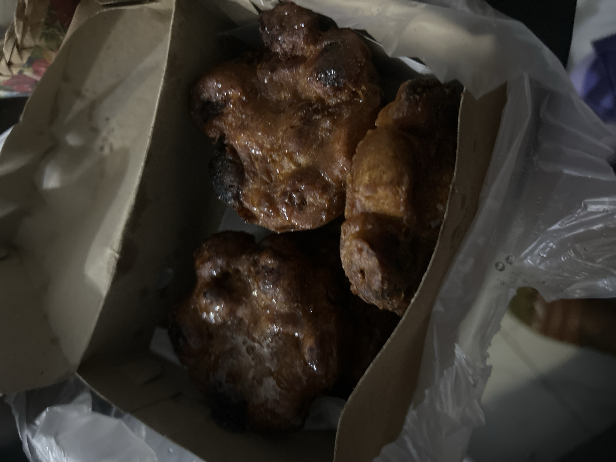 Three deep-fried pastries, possibly *pisang goreng* (fried bananas) or *molen pisang* (banana fritters), are nestled inside a brown paper bag. The pastries are golden brown and appear glazed, with some darker, caramelized areas indicating a crisp exterior. They are irregularly shaped, suggesting they are handmade. The bag is partially open, revealing the treats within, and a crinkled plastic bag is visible in the foreground, possibly used to line the paper bag or as an outer wrapper. The scene is dimly lit, suggesting it might be evening or indoors. The image offers no explicit cues about the time of day or weather, but the context of Sukawati, Indonesia, implies a tropical climate. No people are visible, and there is no visible text. The overall impression is one of freshly prepared, tempting fried pastries.