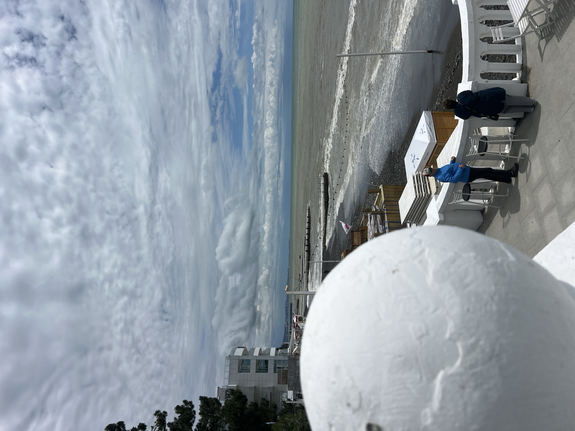 A seaside view in Sochi, Russia, is captured from an elevated position, partially obscured by a prominent white spherical object in the foreground. A white balustrade borders a patio, with two individuals visible: one in a blue jacket and another in a darker jacket observing the scene. Tables and chairs are arranged nearby. The seascape features a shoreline with waves meeting the land, and a sky filled with varied cloud formations. In the distance, buildings are visible near the water's edge.