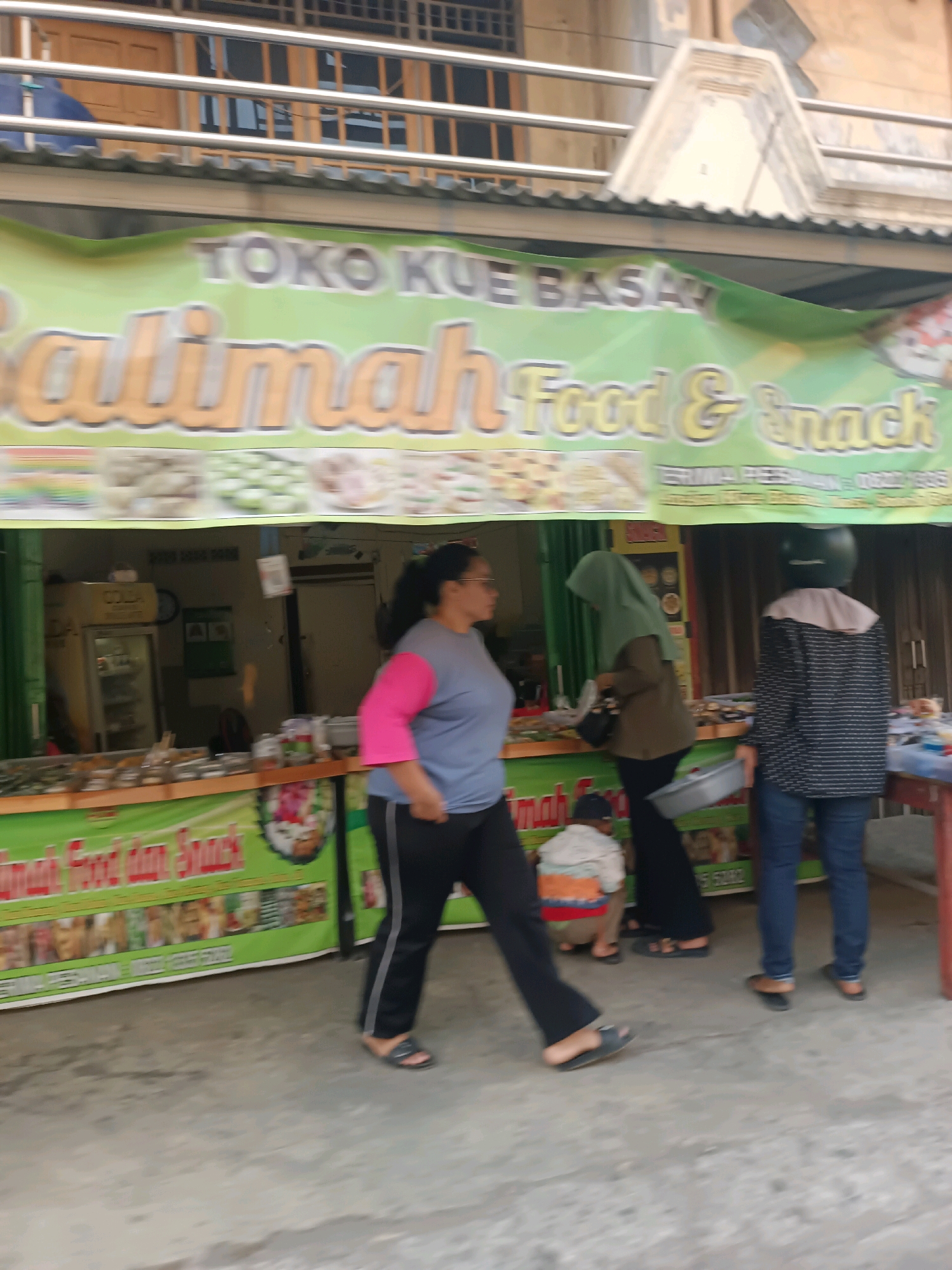 A food stall named "Salimah Food & Snack" is depicted in Karawang, Indonesia, with a green banner displaying the name in large orange letters. The stall appears to be on a sidewalk next to a building with a multi-story structure above. Several people are visible: a woman in a blue shirt with pink sleeves and black pants is walking past the stall. Another woman wearing a green hijab and black pants is standing at the counter. A person in a black and white patterned shirt and blue jeans stands to the right, facing away from the camera. A young child is crouching in front of the stall. Displayed on the counter are various food items.