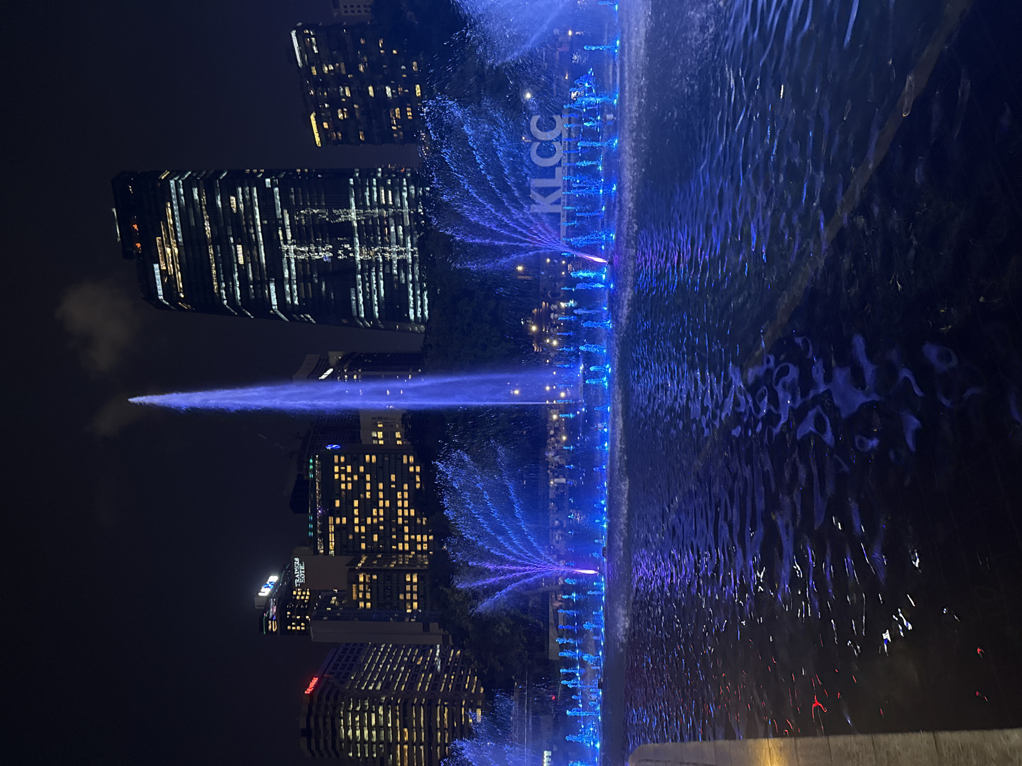 The image shows a nighttime view of the KLCC (Kuala Lumpur City Centre) fountain and surrounding skyscrapers in Kuala Lumpur, Malaysia. The fountain is illuminated with blue lights, and water jets are visible. Reflections of the city lights and the "KLCC" sign are visible in the water. In the background are several skyscrapers with lighted windows. The overall scene conveys a sense of urban activity and nightlife.