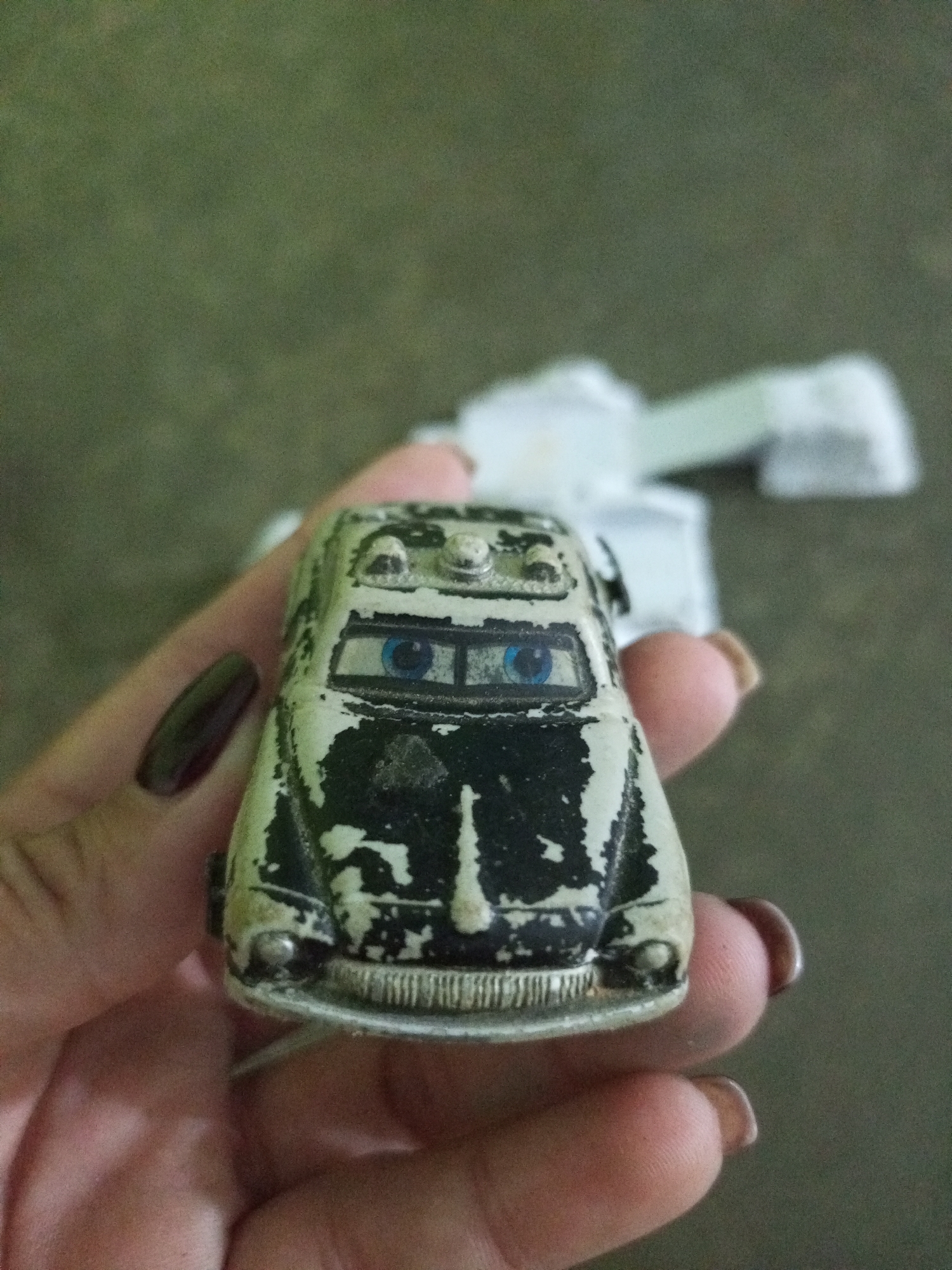 A close-up shot captures a small, weathered toy car with a black and white distressed paint job. The car has large, blue cartoon-like eyes, and two lights on its roof. A hand with dark-colored fingernails holds the toy car. The background is a blurry, dark green surface, possibly a table or ground. There are no visible indicators of the location being in La Tagua, Venezuela.