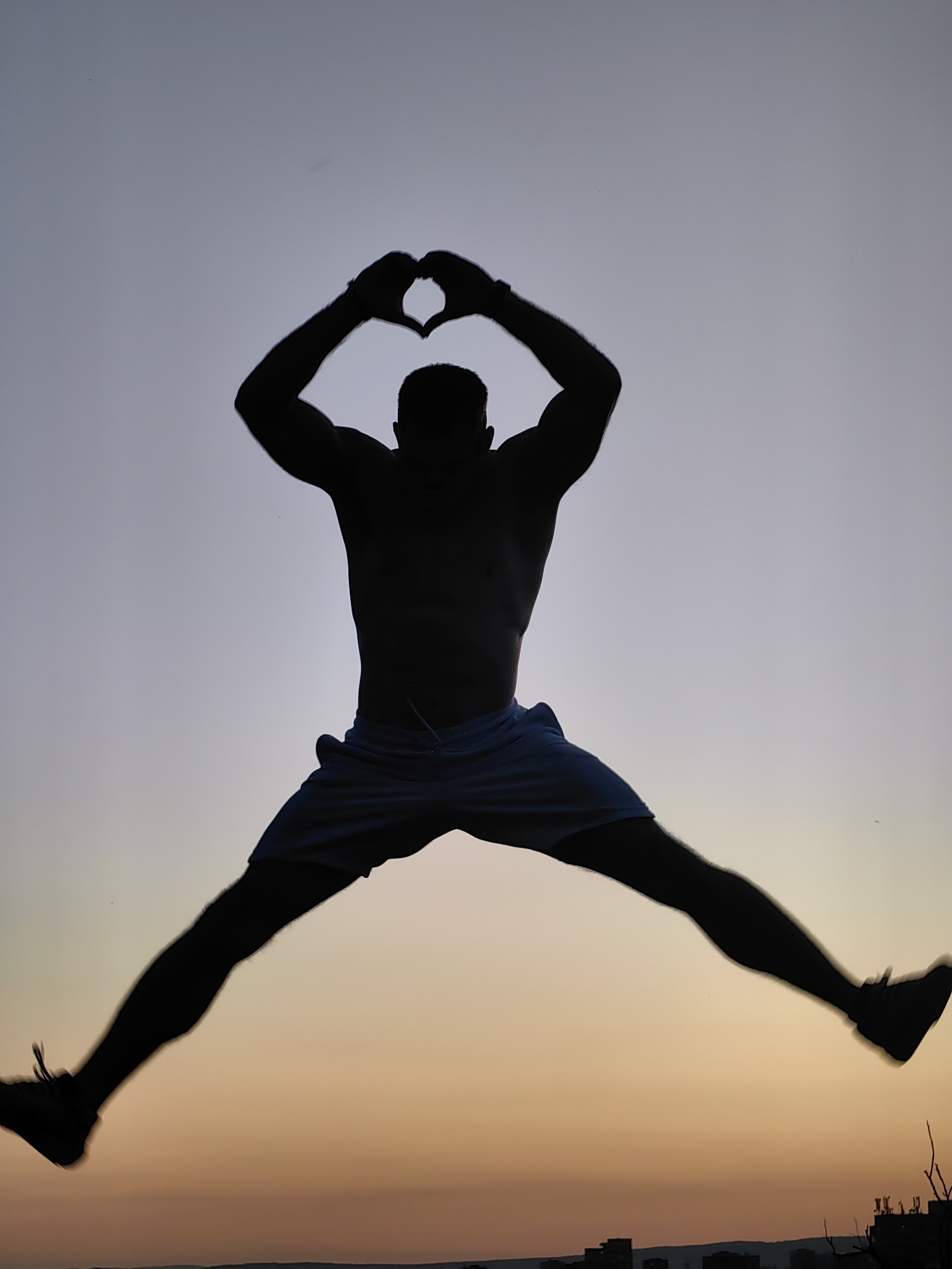 A silhouette of a shirtless man in mid-air, legs spread wide and arms raised to form a heart shape above his head, is captured against a gradient sky transitioning from pale blue to warm orange. The man is wearing light-colored shorts. The image appears to be taken at dusk or dawn, with a blurry cityscape visible along the bottom horizon. The overall impression is one of freedom and joy. The specific location within the city of Varna, Bulgaria, cannot be determined from the image alone.