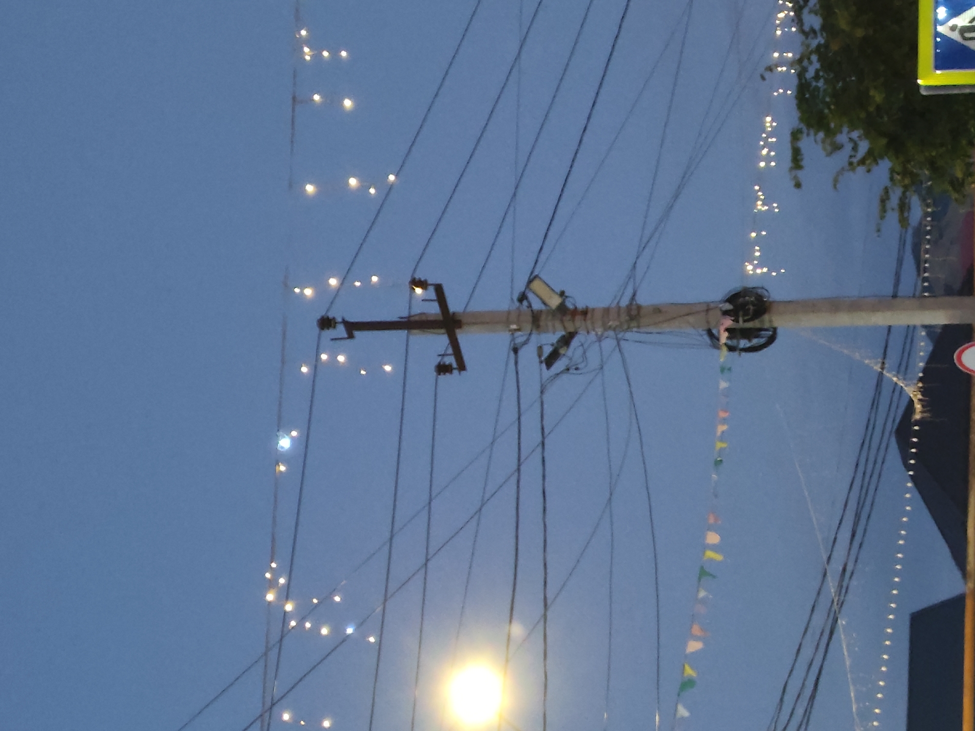 The image shows a street scene at dusk or night in Leninkent, Russia, dominated by a utility pole and overhead wires. The sky is a deep blue, suggesting twilight or a clear night. Strung between wires are numerous small, glowing lights, likely decorative string lights. There are also triangular colorful flags strung along a wire lower in the frame. A bright, round light source, possibly a streetlamp, is visible on the left side of the image, casting a warm glow. The utility pole itself is thick and has several horizontal crossbars supporting the complex network of dark wires. A floodlight fixture is attached to the pole. In the background, the dark silhouettes of rooftops and trees are visible, with more distant, faint lights suggesting buildings or further decorations. A yellow and blue street sign with a pedestrian symbol is partially visible in the lower right corner. The overall impression is a festive or illuminated street environment.