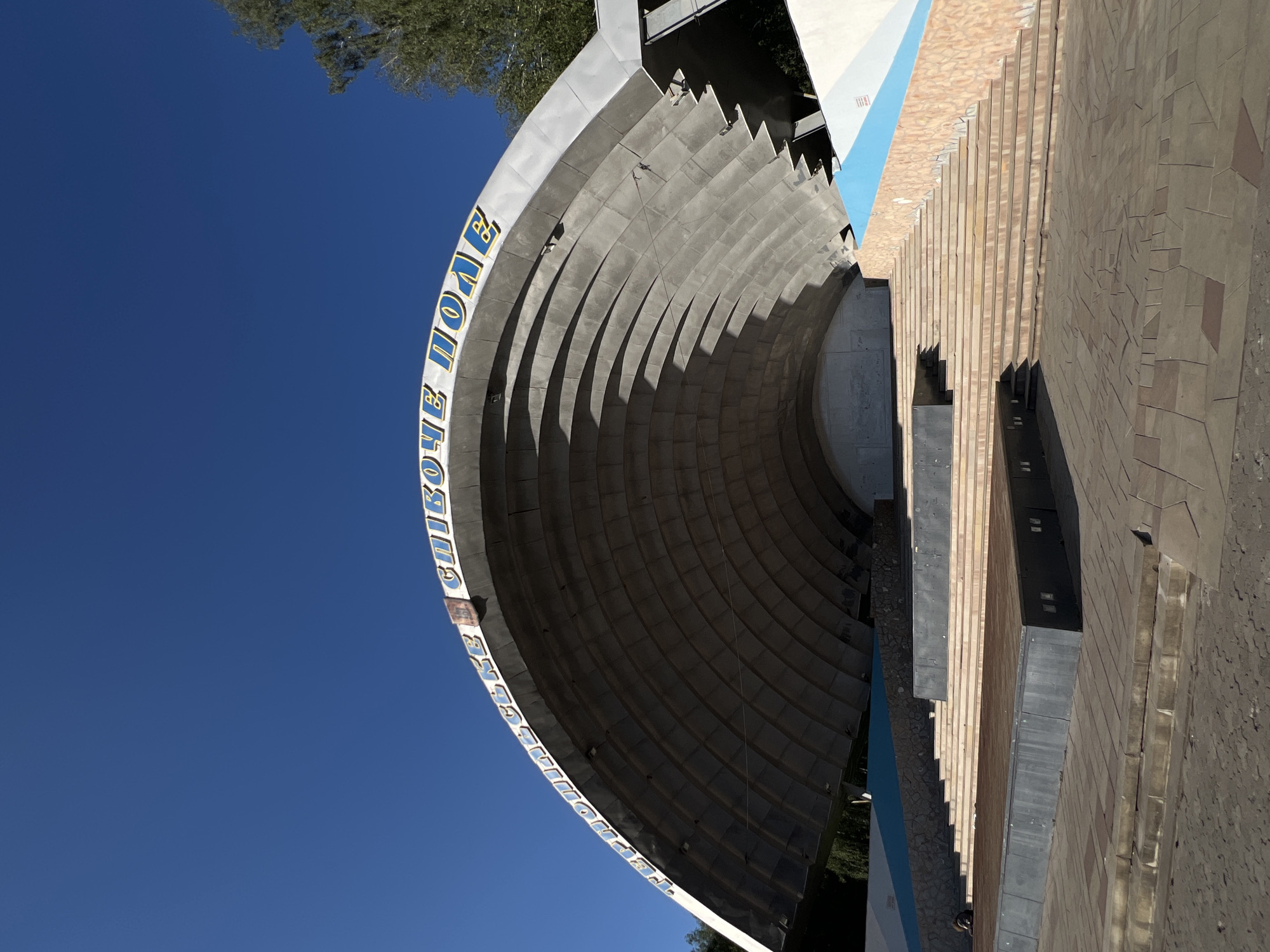 This media file captures an outdoor view of an amphitheater with a curved seating area. The amphitheater's facade features a prominent inscription in blue letters with a white outline. The text, which appears to be in Cyrillic script, reads "СПОРТИВНЕ ПОЛЕ" (Sportive Field). The structure is made of concrete with horizontal ribbed detailing on the seating sections. Adjacent to the amphitheater, there is a stepped area with a warm-toned, tiled surface and a narrow blue strip. The scene is set against a clear, vibrant blue sky, suggesting a sunny day. The presence of trees in the background indicates an outdoor park or recreational area. No people are clearly visible in the frame, and no specific activity or event is depicted. The lighting suggests it is daytime, likely morning or afternoon. The overall impression is one of an open-air venue designed for gatherings or performances, situated in Ternopil, Ukraine.
