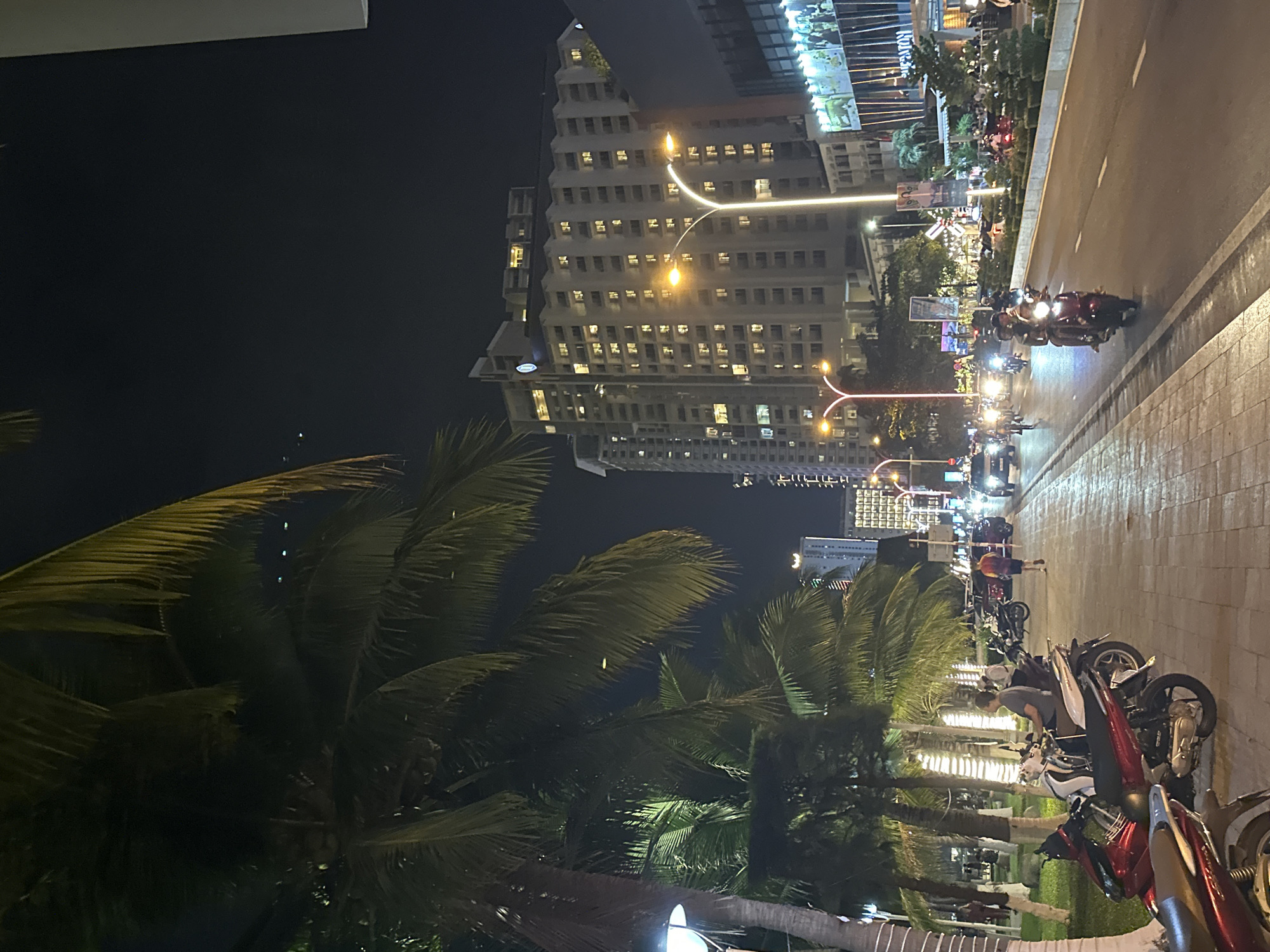 This nighttime photograph depicts a bustling urban street scene in Nha Trang Ward, Vietnam. The setting is illuminated by artificial lights emanating from streetlamps, vehicle headlights, and the windows and facades of buildings, casting a warm glow across the scene.
In the background, a prominent multi-story building, likely a hotel or apartment complex, features numerous illuminated windows, suggesting ongoing activity. Further along the street, other buildings are visible, some with lit signage, including partially visible text that appears to read "KONGA."
The street itself, positioned on the right, is a wide paved road with white lane markings. It is populated by numerous motorbikes (scooters), a common mode of transport in Vietnam. Some motorbikes are in motion, their headlights and taillights creating trails of light, while many others are parked along the spacious sidewalk. In the mid-ground, a person is seen bending over a parked motorbike. Other individuals are scattered throughout the scene, either riding or standing near their vehicles, contributing to the vibrant atmosphere.
Lush palm trees are a significant feature of the landscape, framing the left side of the image and lining portions of the street. Their dark fronds are highlighted by the surrounding artificial lights. The road surface appears damp and reflective, possibly due to recent rain or high humidity. The overall impression is one of a lively city evening.