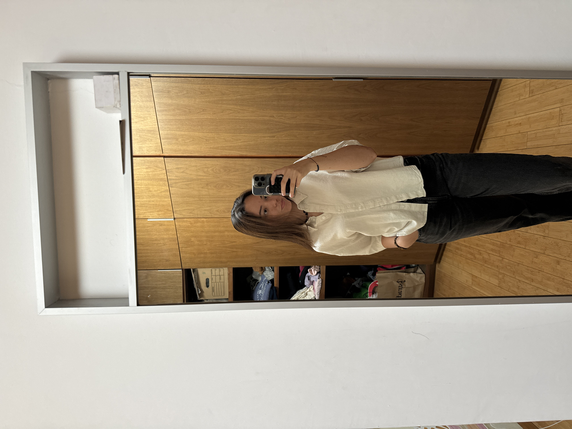 A full-length mirror selfie shows a person, likely female, standing indoors. The image is taken in a rectangular mirror with a light grey frame, positioned against a plain white wall. The person is wearing a light, possibly off-white, collared long-sleeved shirt and dark pants, which appear to be jeans. They hold a dark-colored smartphone, possibly an iPhone, in their right hand to capture the reflection and are looking directly into the camera. A dark bracelet is visible on their left wrist.
Reflected behind the subject is a wooden cabinet or wardrobe with a warm, natural wood finish, featuring multiple panels or doors, some with horizontal metal handles. An open section of the cabinet below the person's right arm reveals shelves containing various items, including what appears to be folded clothing and a brown paper bag. Faint, reversed text, possibly "bepq" or "qpeb", is partially visible on the bag. The floor reflected in the mirror consists of light-colored wooden planks. A small, rectangular white object is attached to the top-left inner corner of the mirror frame. The scene is well-lit, suggesting daytime. The person maintains a neutral expression while posing for the photo in San Bernardino Tlaxcalancingo, Mexico.