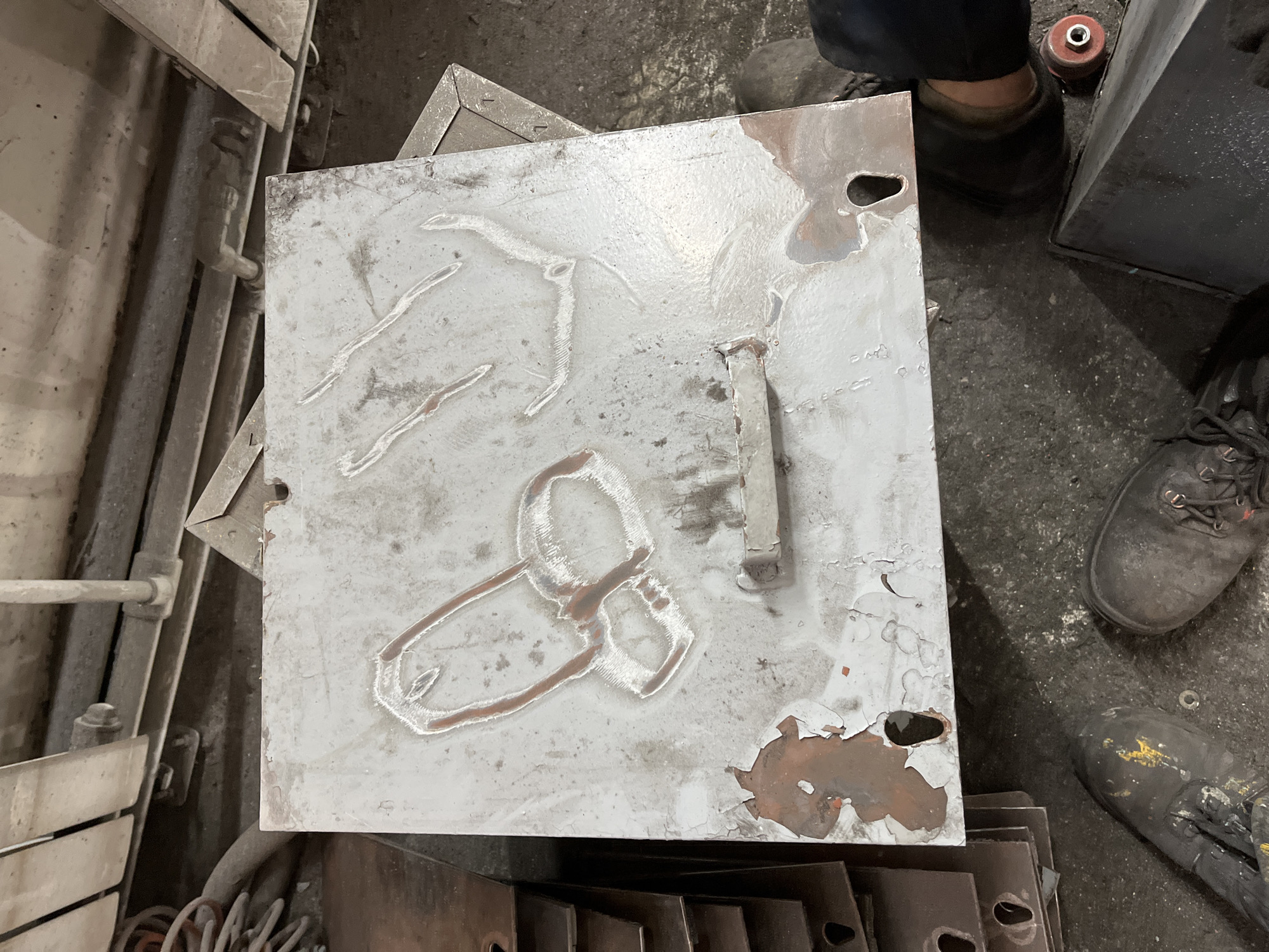 This media file presents an overhead view of a square metal plate within what appears to be an industrial workshop in Chaikovsky, Russia. The central subject is a large, greyish-white metal sheet, possibly steel, whose paint coating is peeling extensively, revealing patches of reddish-brown rust underneath. Etched or welded onto its surface are several distinct designs: one resembles a stylized animal, possibly a deer or antelope, while another prominently depicts a phallic symbol. The plate also features two irregularly shaped holes in its upper right and lower right corners.
Beneath this main plate, several smaller, darker metal sheets with similar cutouts are stacked. The surrounding environment includes various industrial elements: metal pipes and a section of a large machine or structure on the left, and a dark, angular object on the right. The floor is a dark, rough surface, likely concrete or asphalt.
Parts of two individuals are visible in the frame, specifically their legs and feet, clad in sturdy, dark work boots, indicating their presence in this industrial setting. One boot on the bottom right has noticeable yellow paint marks. A small red cylindrical object is also visible near one of the feet. The image suggests a moment during metalwork, a piece being processed, or perhaps a display of informal artistic expression on industrial material. No discernible text is visible.