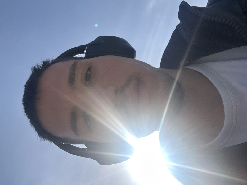 The image displays a low-angle, sun-drenched selfie of a male individual outdoors. The subject has short, dark hair and light facial stubble, directly facing the camera with a neutral expression. He is wearing dark, over-ear headphones, a white crew-neck t-shirt, and a dark jacket. Strong sunlight emanates from the lower-left quadrant, creating significant lens flare that partially obscures the left side of the subject's face and upper torso. The background is a clear blue sky, with no discernible environmental features beyond it. Visual analysis of the image does not provide sufficient detail to ascertain a specific location within the city of Varna, Bulgaria.