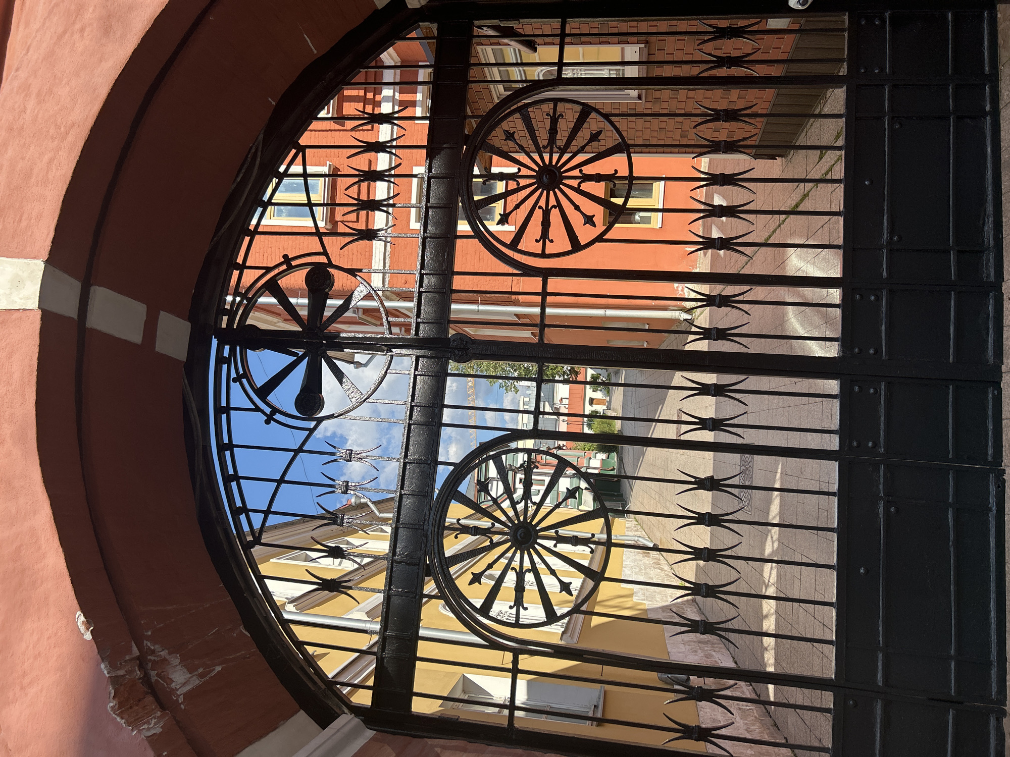 An arched gateway frames a narrow courtyard or alleyway between two buildings. The gateway is constructed of black wrought iron, featuring a large, ornate circular motif resembling a spoked wheel with decorative elements on its left and right sides. The gate itself is also black iron and appears to be closed. The buildings flanking the courtyard are painted in bright colors: one is yellow with white trim and windows, and the other is a vibrant orange-red brick. The ground of the courtyard is paved with light-colored square tiles. The sky above is blue with scattered white clouds. Sunlight casts distinct shadows from the ironwork onto the courtyard pavement and the buildings. The scene appears to be outdoors in a residential or historic area.