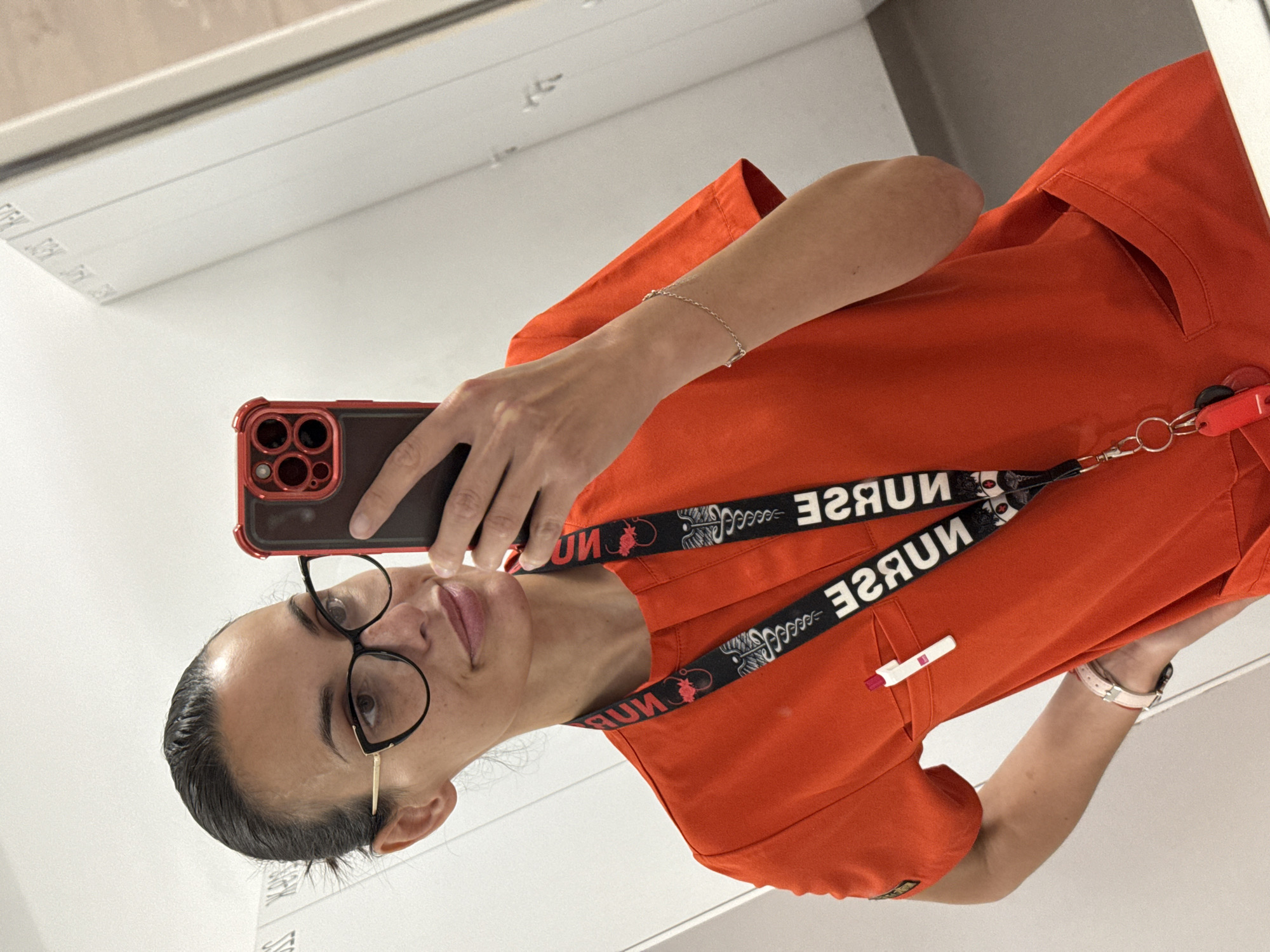 The image displays a person taking a mirror selfie, presented rotated 90 degrees counter-clockwise. The subject, a female with dark hair pulled back and wearing black-rimmed glasses with gold-colored arms, has a neutral expression with a slight smile. She is dressed in vibrant orange medical scrubs.
Around her neck, a black lanyard is visible, bearing the mirrored text "NURSE" repeated, along with a medical symbol resembling a caduceus, indicative of a healthcare profession. A white pen with a red tip is clipped into a pocket on her scrubs.
She holds a smartphone, an iPhone with a distinct red protective case and multiple camera lenses, in her right hand to capture the reflection. A delicate, thin silver-colored chain bracelet is on her right wrist, and a watch with a light-colored strap (possibly pink or beige) is on her left. The background visible in the mirror's reflection is a plain, light-colored indoor environment, consistent with a medical facility. The scene is set in Pleven, Bulgaria. No distinct time of day or weather conditions are discernible from the indoor setting.