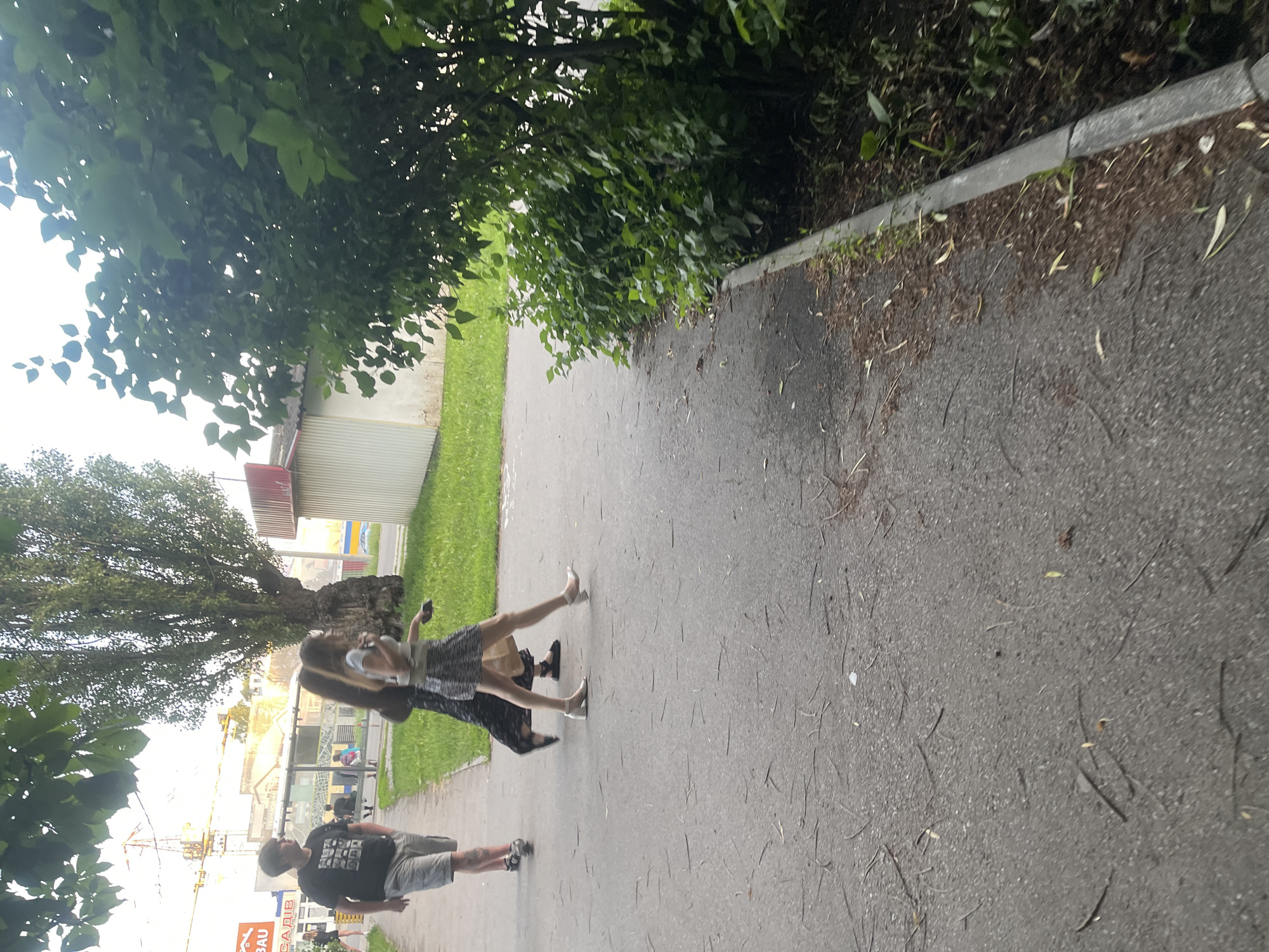 A low-angle medium shot captures three individuals walking on a paved walkway adjacent to a grassy area and dense foliage. In the foreground, a person with dark hair and a black graphic t-shirt and grey shorts walks from left to right. Behind them, two women walk in the same direction. The woman in the middle has dark hair and wears a long, dark dress. The woman in front has long brown hair and wears a short, patterned skirt and a white top. She is wearing heels, and the other woman is wearing flat sandals. In the background, a tall tree with a thick trunk stands to the left, with a building and a crane visible behind it. To the right, a white building with a red roof is partially obscured by a large bush with green leaves. The ground is covered in small twigs and leaves.