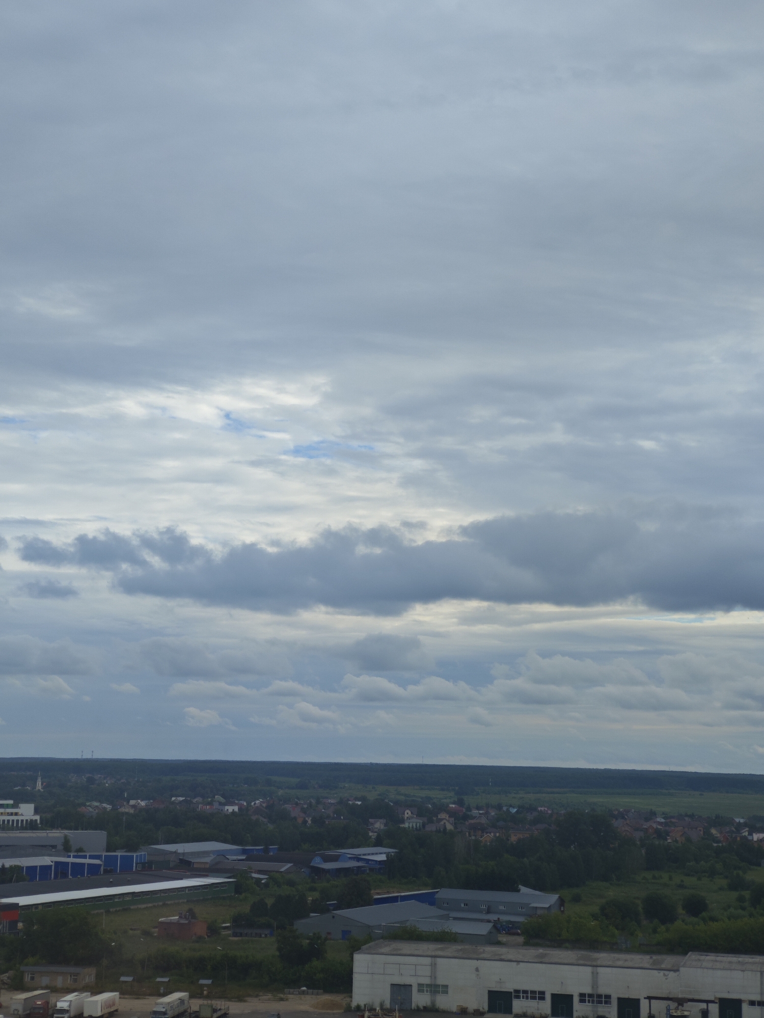 This is an aerial view of an industrial area and a residential town under a cloudy sky. The scene depicts a landscape with buildings, trucks, and trees. The weather appears overcast, suggesting a possibly cool or damp day. The time of day is not definitively indicated, but the lighting suggests it could be daytime. The location is identified as Chekhov, Russia, which is supported by the presence of typical Russian architecture in the residential area and the overall landscape. There is no discernible activity or event taking place; it's a static view of a developed area. Some of the buildings are large industrial structures with flat roofs and loading docks. Smaller residential houses with red and brown roofs are visible further in the distance, interspersed with lush green trees. Several trucks, including semi-trucks and box trucks, are parked in what appears to be a lot or loading area in the foreground, indicating ongoing logistics or commerce. The sky is filled with layered clouds, ranging from light gray to darker, more ominous tones, with hints of blue sky peeking through in some areas. The overall impression is one of a functional, somewhat gray industrial and residential landscape under a moody sky. No text is visible in the image.