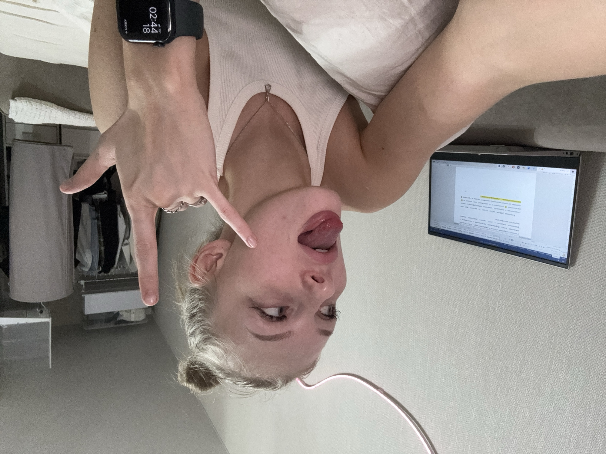 In a bedroom environment, a young woman with blonde hair styled in a bun is shown in a selfie. She is wearing a white tank top and a silver necklace. Her tongue is sticking out, and she is making a rock-and-roll hand gesture with her right hand, while her left hand is visible near her face. A laptop displaying text is positioned to her left, and a closet filled with clothes is visible in the background. The room appears to be in an apartment, possibly in the city of Perm, Russia, given the context provided, although no definitive external location markers are present in the image itself. The lighting suggests it is daytime or the room is well-lit.