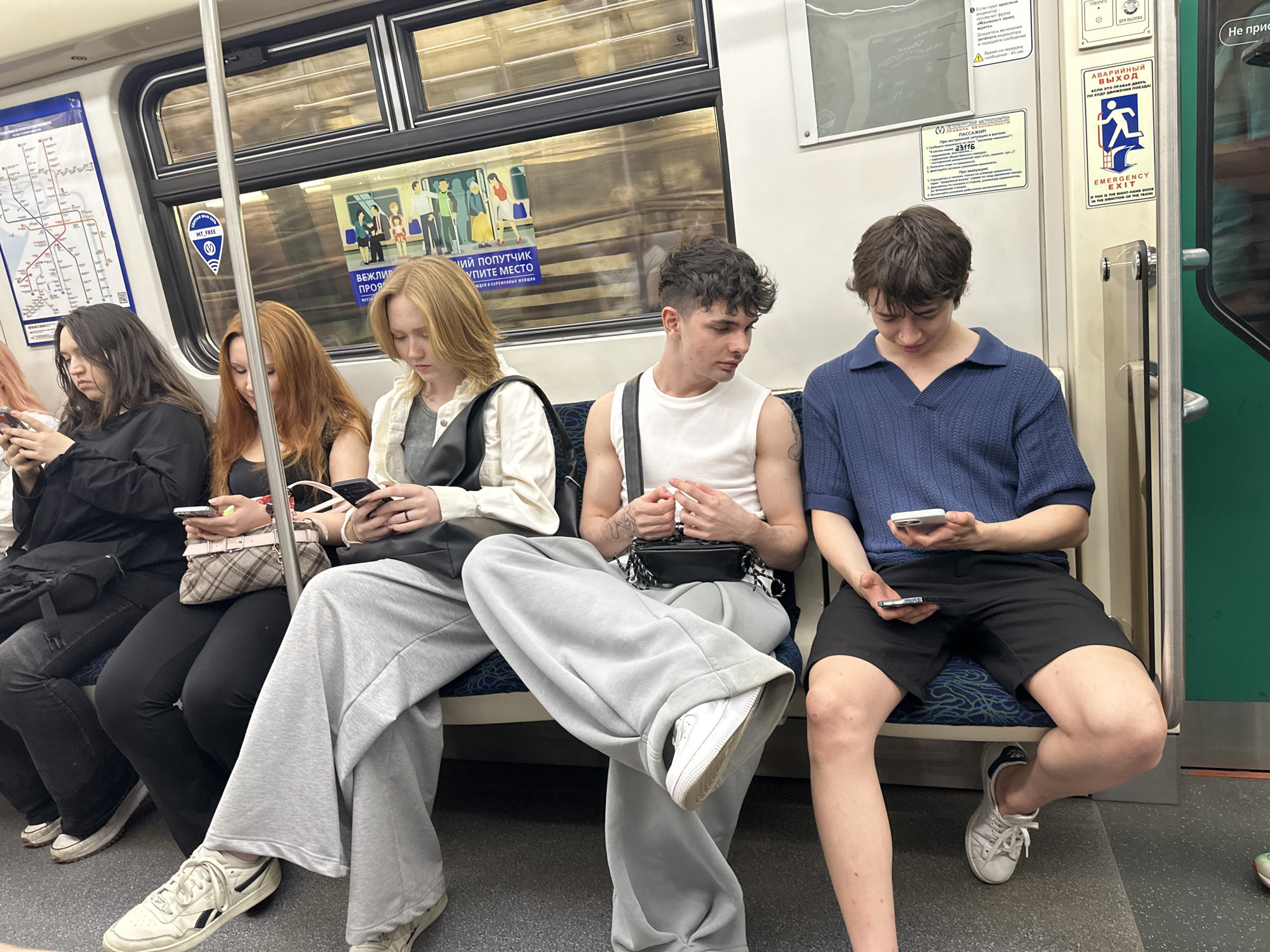 The photograph, taken in Saint Petersburg, Russia, depicts five young adults seated side-by-side on a subway train. The setting is within the train car, evidenced by the presence of a map, various signage, windows, and metal fixtures consistent with subway interiors. The individuals are all occupied with their phones, and wearing contemporary casual clothing. They appear to be of mixed ethnicities. The lighting is uniform, suggesting overhead fluorescent illumination. The overall atmosphere is typical of an urban transit environment.