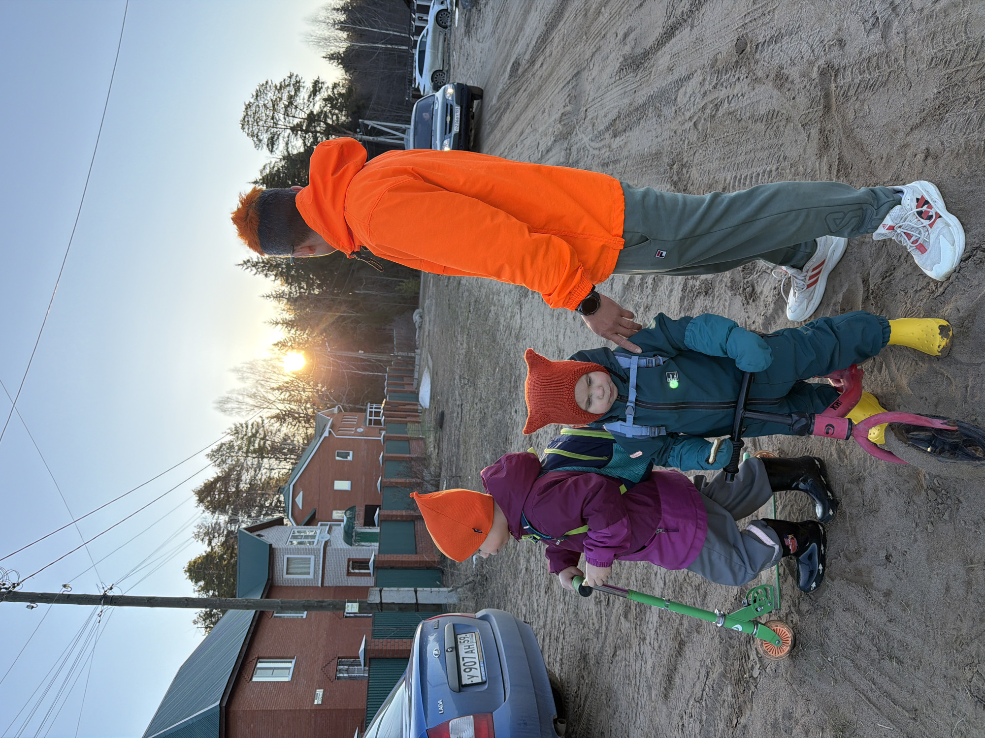 Outside in a sandy area of Chaikovsky, Russia, a man in an orange jacket and green pants stands near two children on scooters. The children wear orange hats and winter clothes. One child is riding a green scooter, while the other is on a pink one. A blue Lada car with the license plate "Y 907 AH 59" is parked nearby. In the background are brick houses and bare trees, with the setting sun visible.