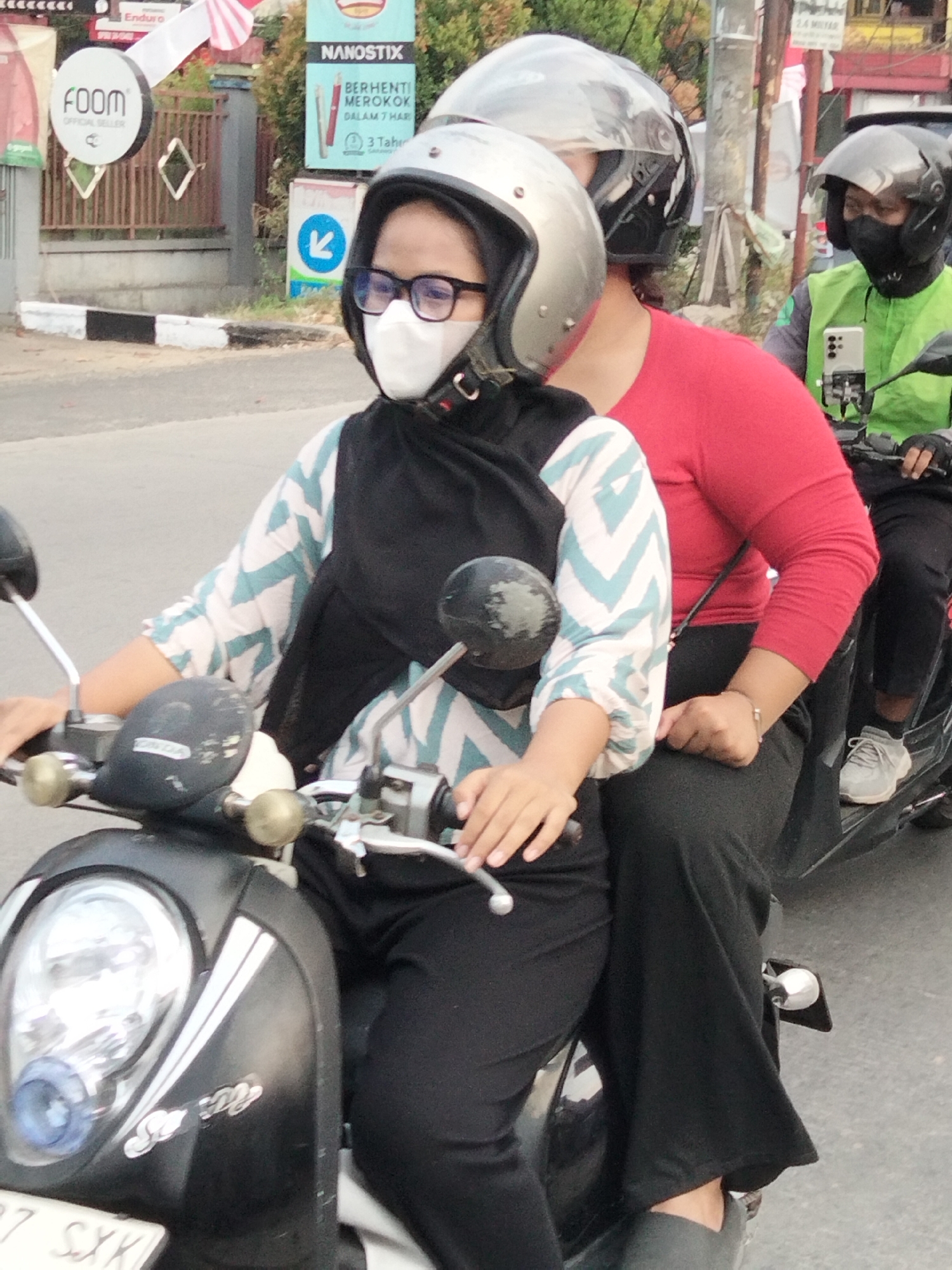 Two individuals are riding a black scooter on a paved road in what appears to be an urban or suburban area of South Tangerang, Indonesia. The front passenger, wearing a black hijab, a white face mask, glasses, and a patterned blue and white shirt, is seated on the scooter. Behind her, the driver is wearing a red long-sleeved shirt and a black hijab. A third person, likely the driver of the scooter, is partially visible behind the second rider, wearing a bright green vest over a grey shirt, a dark helmet, and a black face mask, and is operating the scooter. The scooter has a prominent headlight and chrome accents. In the background, there are signs and a fence, suggesting a commercial or residential area. One sign displays "NANOTIX" and text in Indonesian, another shows a circular logo with "FOOM". The weather appears to be overcast.