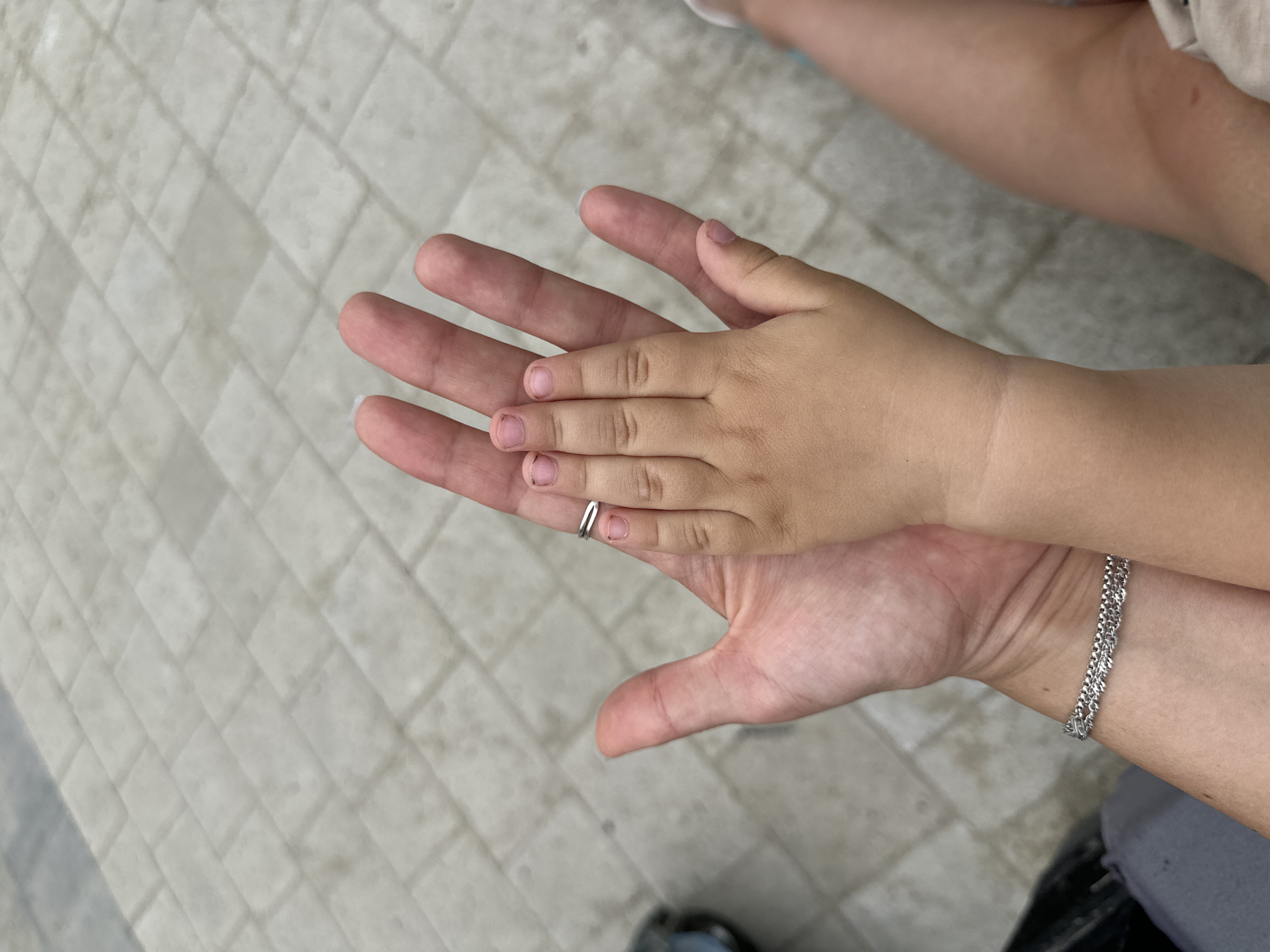Two hands, one significantly larger than the other, are held palm-up against a light grey paved surface. The smaller hand, presumably a child's, rests on the palm of the larger hand, fingers extended and splayed. Both hands exhibit visible fingernails, some with nail polish. A thin silver ring is visible on one finger of the larger hand, and a delicate silver bracelet adorns the wrist of the larger hand. The background is a textured, light grey paving pattern, resembling interlocking bricks. The scene is captured from a top-down perspective. The location in Malinovka, Russia, is not discernible from the provided image.