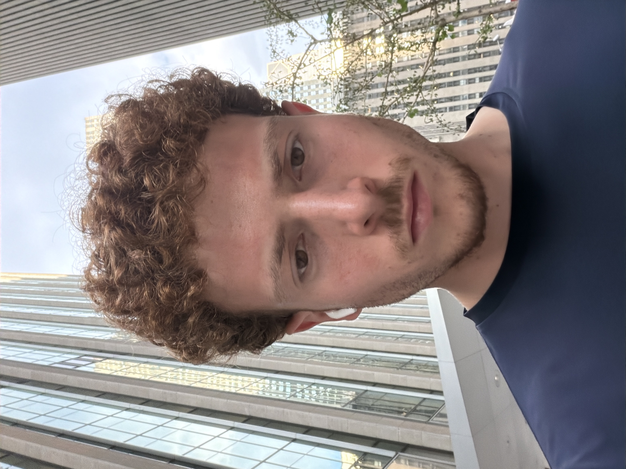 A man with curly brown hair, light skin, and a short brown beard takes a selfie against an urban backdrop. He wears a navy blue shirt and has a white earbud in his ear. The background features buildings with numerous windows, suggestive of a city environment, likely in New York, United States of America.
