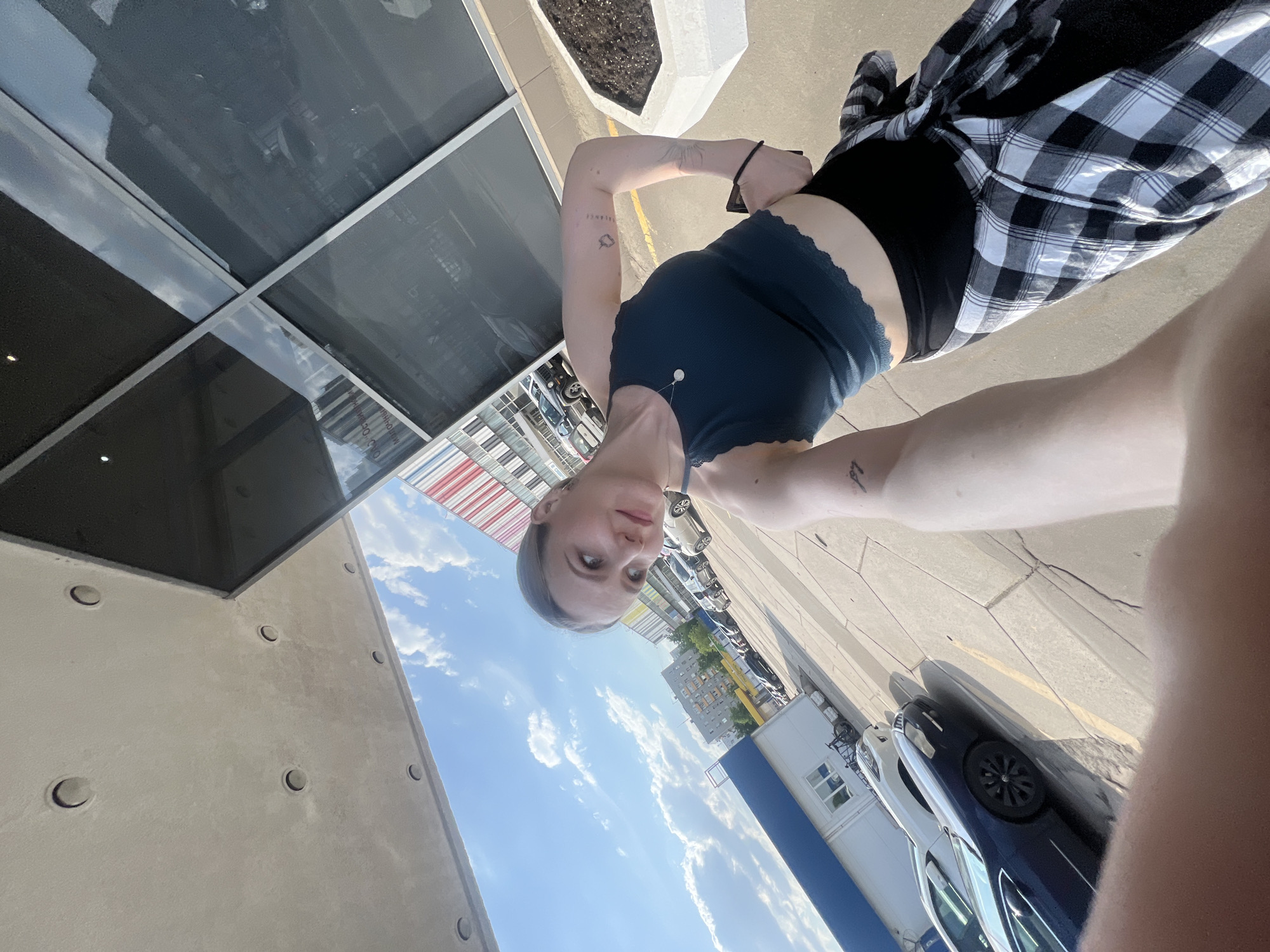 A young woman is taking a selfie outside a building with large windows that reflect the sky. The building appears to be a commercial or office building. The woman is wearing a dark blue tank top, black shorts, and a black-and-white plaid shirt tied around her waist. Tattoos are visible on her arms. The location seems to be an urban area with cars parked on the street. The sky is clear with scattered clouds. There is a flower pot next to the building. The city is not visible in the image.