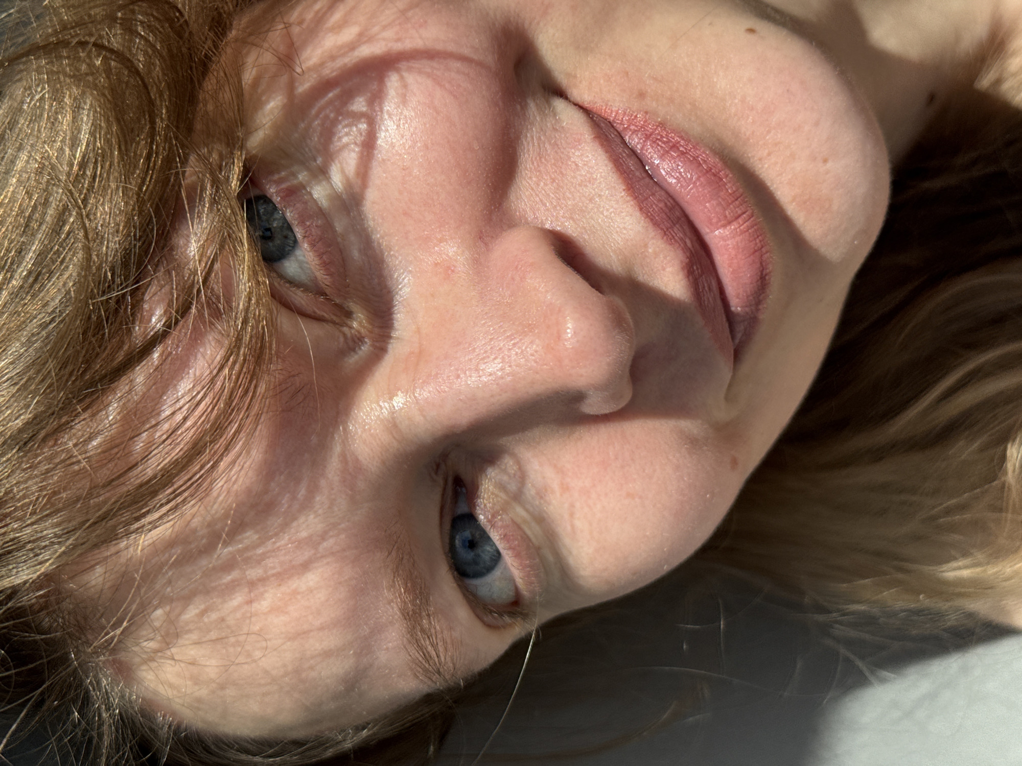 The image is a close-up shot of a person's face, likely female, with light skin, light brown hair, and blue eyes. The face is tilted towards the camera. The lighting is bright and creates strong shadows on the face. The lips are visible and appear to have a nude or light pink lipstick applied. No details suggest the city of Izhevsk, Russia.