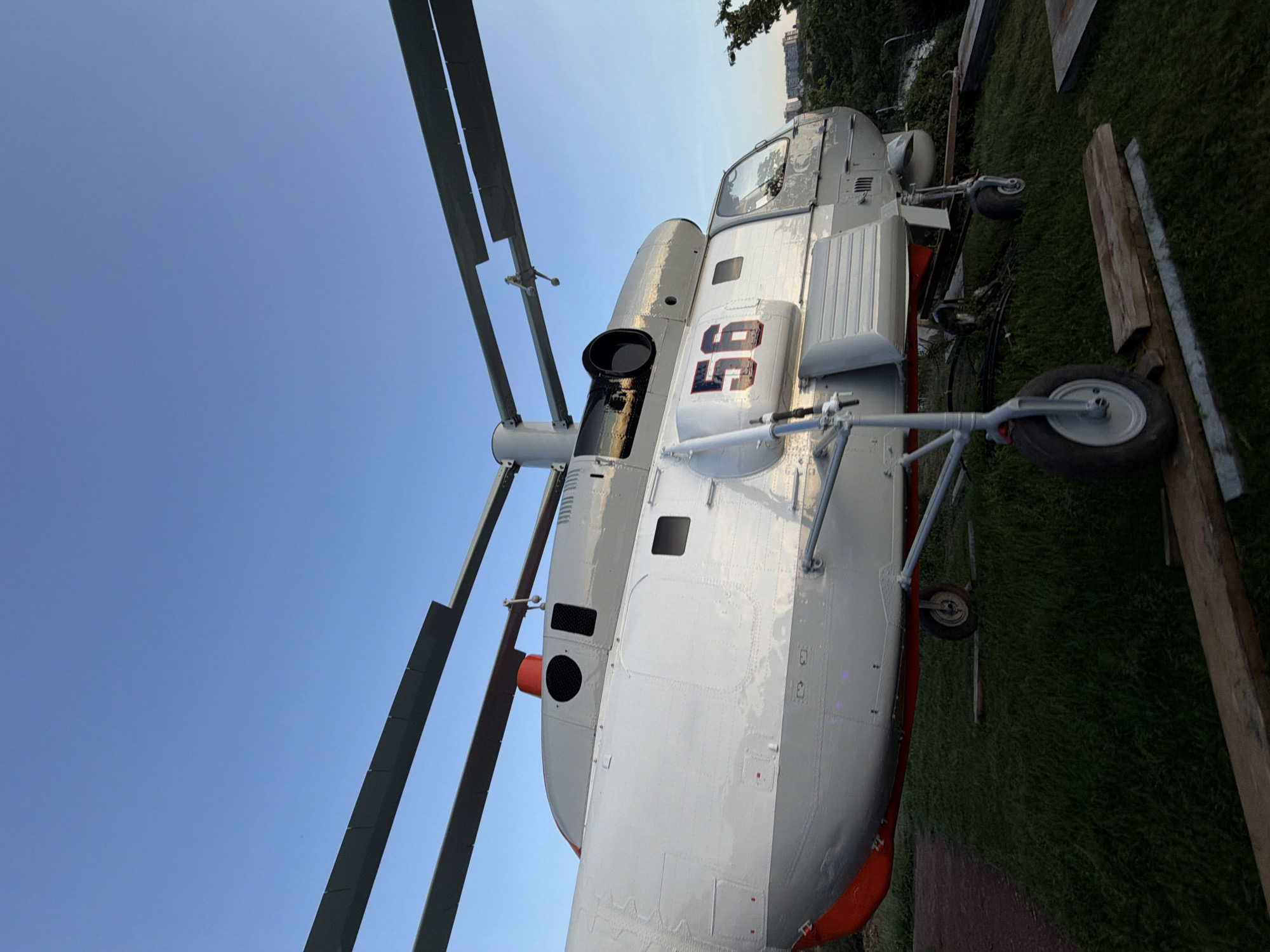 A low-angle, full-shot photograph captures a parked helicopter, identified by the number "56" on its side, against a clear blue sky. The helicopter, painted white and gray with red accents on its lower fuselage, rests on a grassy incline. Its rotor blades are visible, extending horizontally across the top of the frame. The helicopter's landing gear, including front and rear wheels, is also prominent. The scene appears to be an outdoor exhibit or display, possibly at a park or museum. In the background, a distant cityscape with buildings and trees is visible, suggesting an urban or semi-urban setting. The overall lighting suggests it is either early morning or late afternoon. There are no people visible in the image. The location is identified as Kamyshin, Russia.