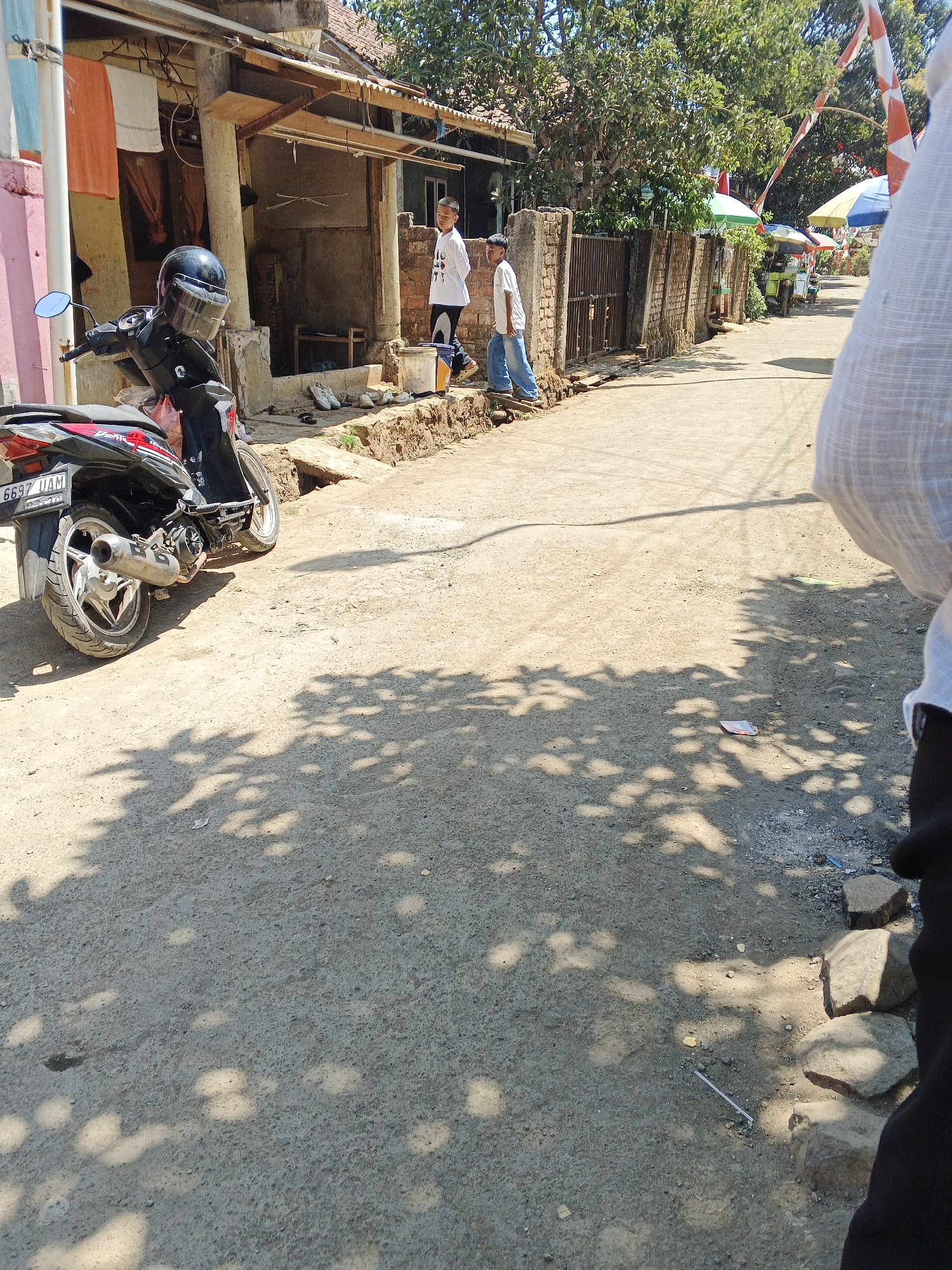 The image captures a street scene in Sukaharja, Indonesia, under bright daylight. A dirt road with scattered rocks and debris runs through the frame, dappled with the shadows of overhead foliage. On the left, a black motorcycle with a red accent and license plate "6697 UAM" is parked. A black helmet rests on the handlebars. Adjacent to the motorcycle is a building with a wooden porch and a corrugated metal roof. Orange and white laundry hangs from a clothesline outside the building.
Further down the street, two young boys are visible. One, wearing a white t-shirt and dark pants, stands near a brick wall. The other boy, in a white t-shirt and blue jeans, is walking away from the viewer. The brick wall continues along the right side of the street, featuring a metal gate. Behind the wall, several colorful umbrellas are set up, suggesting a market or street vendor area. A small green vehicle, possibly a motorcycle rickshaw, is parked behind one of the umbrellas. Indonesian flags, red and white, are strung across the scene, indicating a possible national celebration. The foreground on the right shows a portion of a person's arm and shoulder, clad in a white textured garment, implying the photographer's presence. The overall atmosphere is that of a typical, lively Indonesian village street on a sunny day.