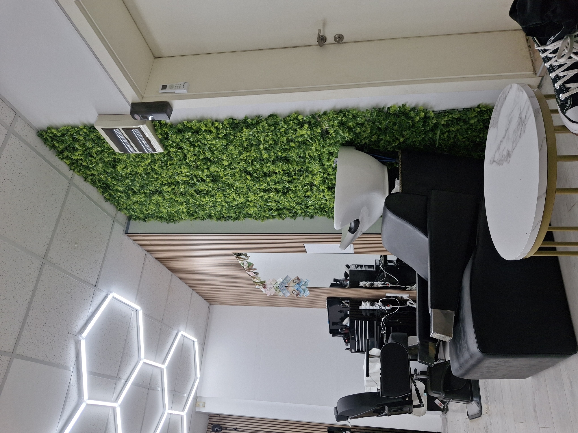 The image depicts the interior of a modern hair salon or barber shop in Sofia, Bulgaria. The establishment features light-colored laminate flooring and white walls, with design elements including a vertical light wood slat accent wall and a floor-to-ceiling faux green plant wall. The ceiling consists of white acoustic tiles, from which a series of interconnected hexagonal LED light fixtures are suspended, providing bright illumination.
In the mid-ground, a black, adjustable hydraulic barber chair is visible, and further back, another similar chair is partially obscured. Adjacent to the faux green wall, a white ceramic hair washing basin with a black base is installed. To the left of the washing station, a black multi-drawer tool chest on wheels, stocked with various salon tools and products, is positioned next to a large wall-mounted mirror. A decorative string of assorted Bulgarian leva banknotes (denominations visible include 2, 5, 10, 20, and 50 leva) is suspended above the mirror.
On the white wall above the faux green wall, a white rectangular air conditioner unit and a black air freshener dispenser are mounted. A white standard door, featuring a silver handle and lock, is visible on the right side of the frame. A remote control unit, labeled "KABAI," is taped to the door frame. In the foreground, a small, round side table with a white marble-patterned top and gold-colored legs stands near a reclining black leather shampoo chair. A pair of black high-top sneakers with white laces are partially visible on the floor in the bottom right corner. No people are present, and no active interactions or actions are depicted in the scene. The Bulgarian currency confirms the location as Bulgaria, specifically a commercial establishment within the city of Sofia.