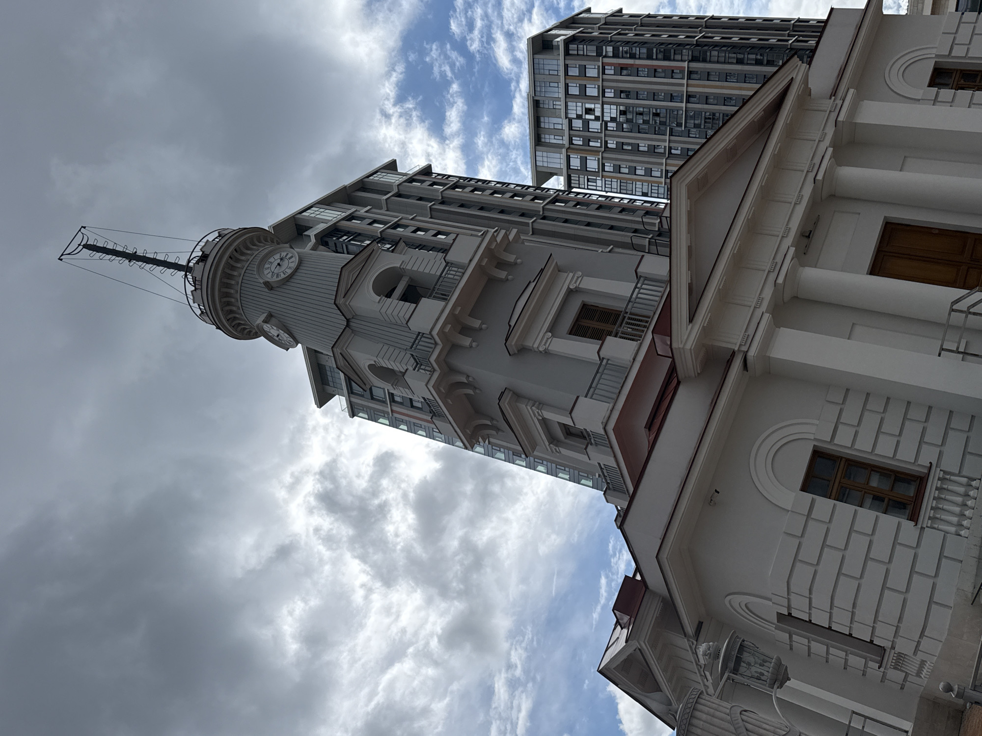 The image shows the historic building of the Ministry of Railways, located at Novo-Basmannaya Street, 2, Moscow, Russia. The building features classical architecture with a white facade, columns, and a prominent clock tower. Behind it, two modern high-rise buildings rise, contrasting sharply with the historical architecture. The sky is cloudy, with patches of blue sky visible. The image is taken from a low angle, emphasizing the height of the buildings. No people are visible in the frame.