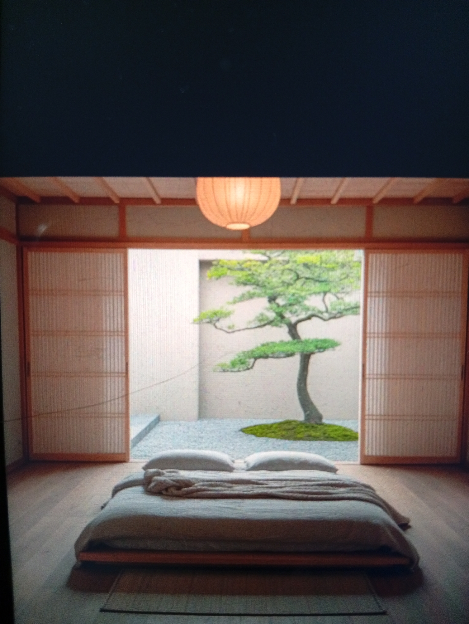 A bedroom with Japanese-inspired decor, featuring a low-platform bed with light-colored bedding and two pillows. A blanket is draped on the bed. Behind the bed, sliding doors made of translucent paper and wood open to a small courtyard garden with a single, sculpted tree growing on a mossy patch. Gravel covers the ground of the garden. A paper lantern hangs from the ceiling of the room. The location of Dutsen Alhaji, Nigeria, is not evident in the image.
